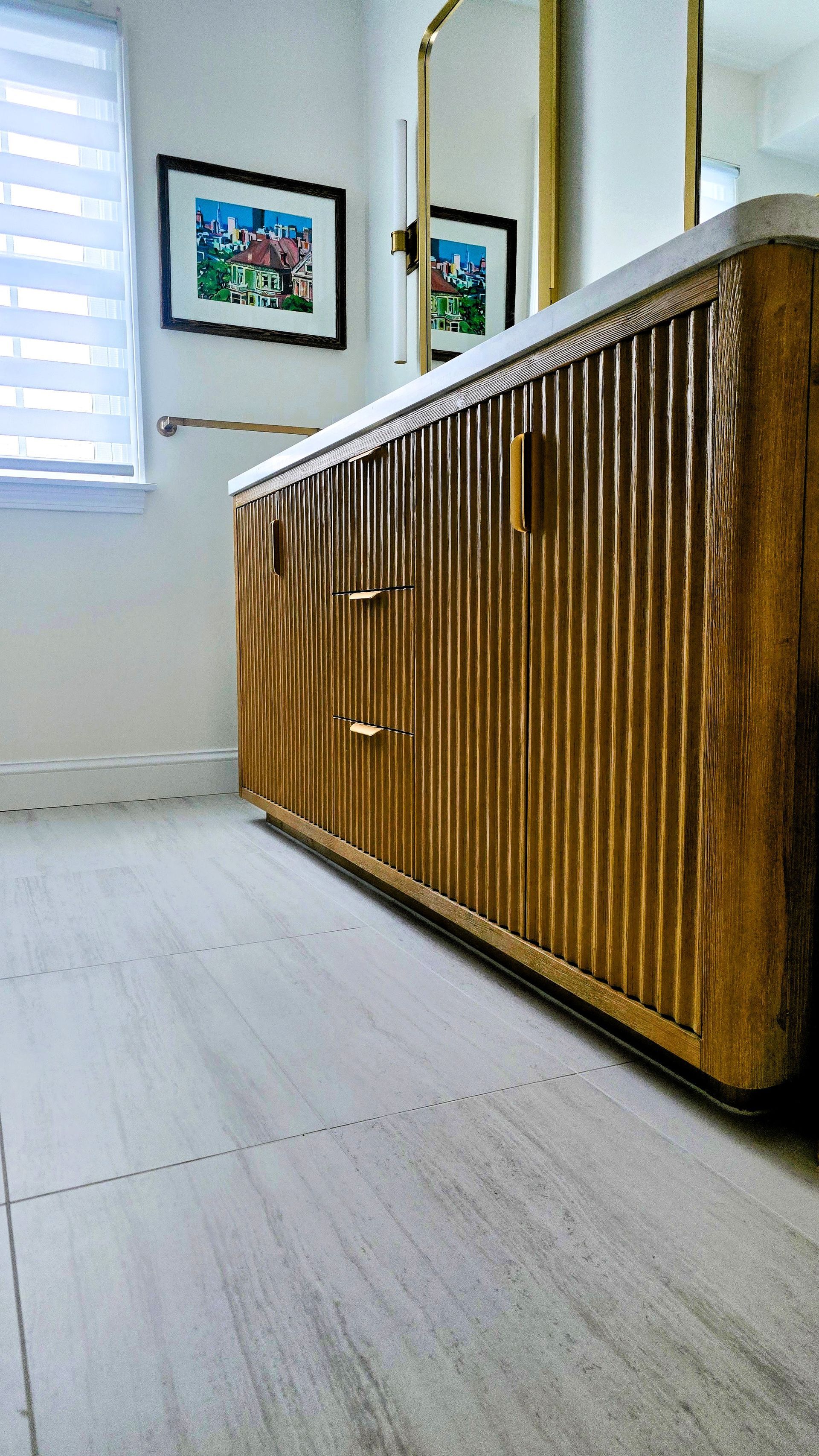 A vanity with a slatted wood finish and gold handles stands in a white bathroom in Cromwell, CT next to a window and a wall mirror.