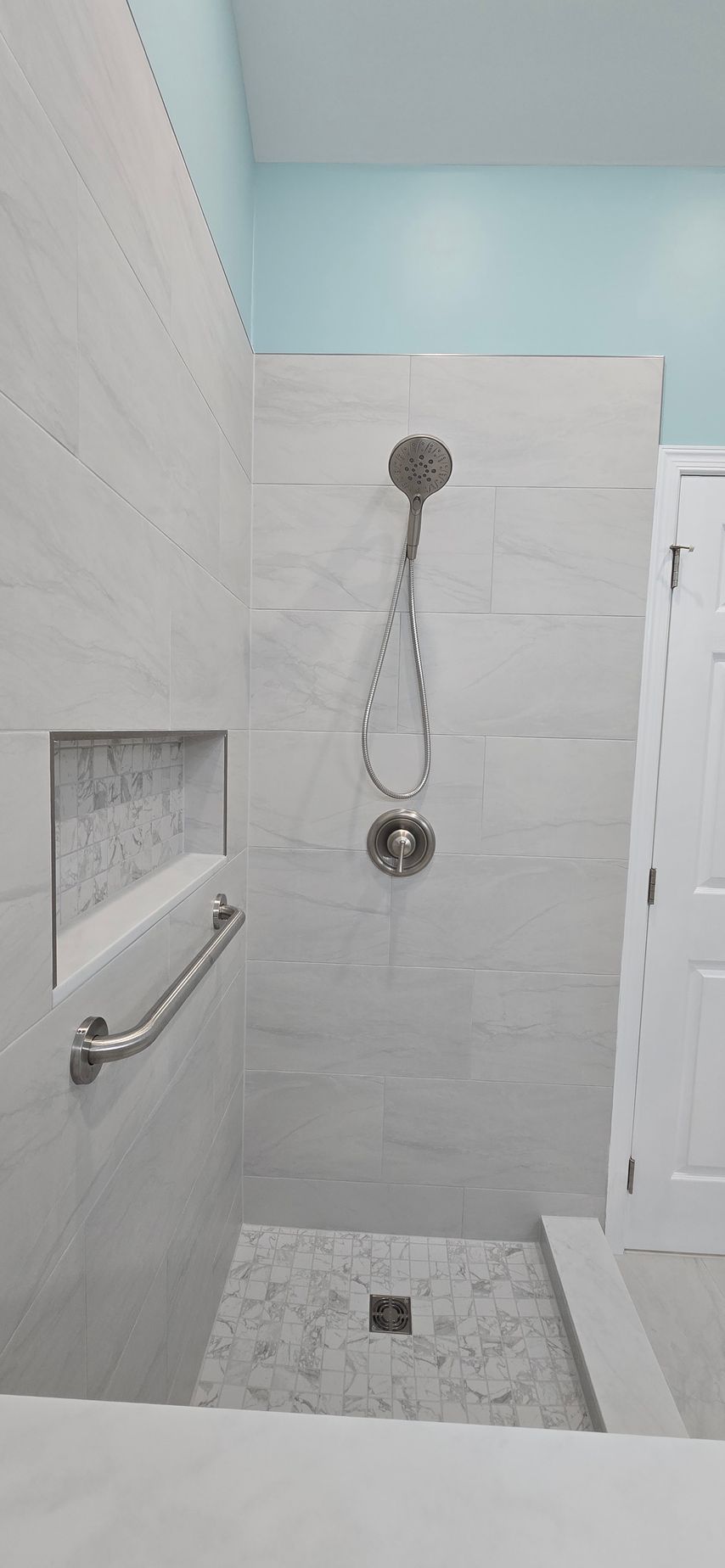 A tiled shower with a hand shower and grab bar. Light gray tiles and a light blue wall.
