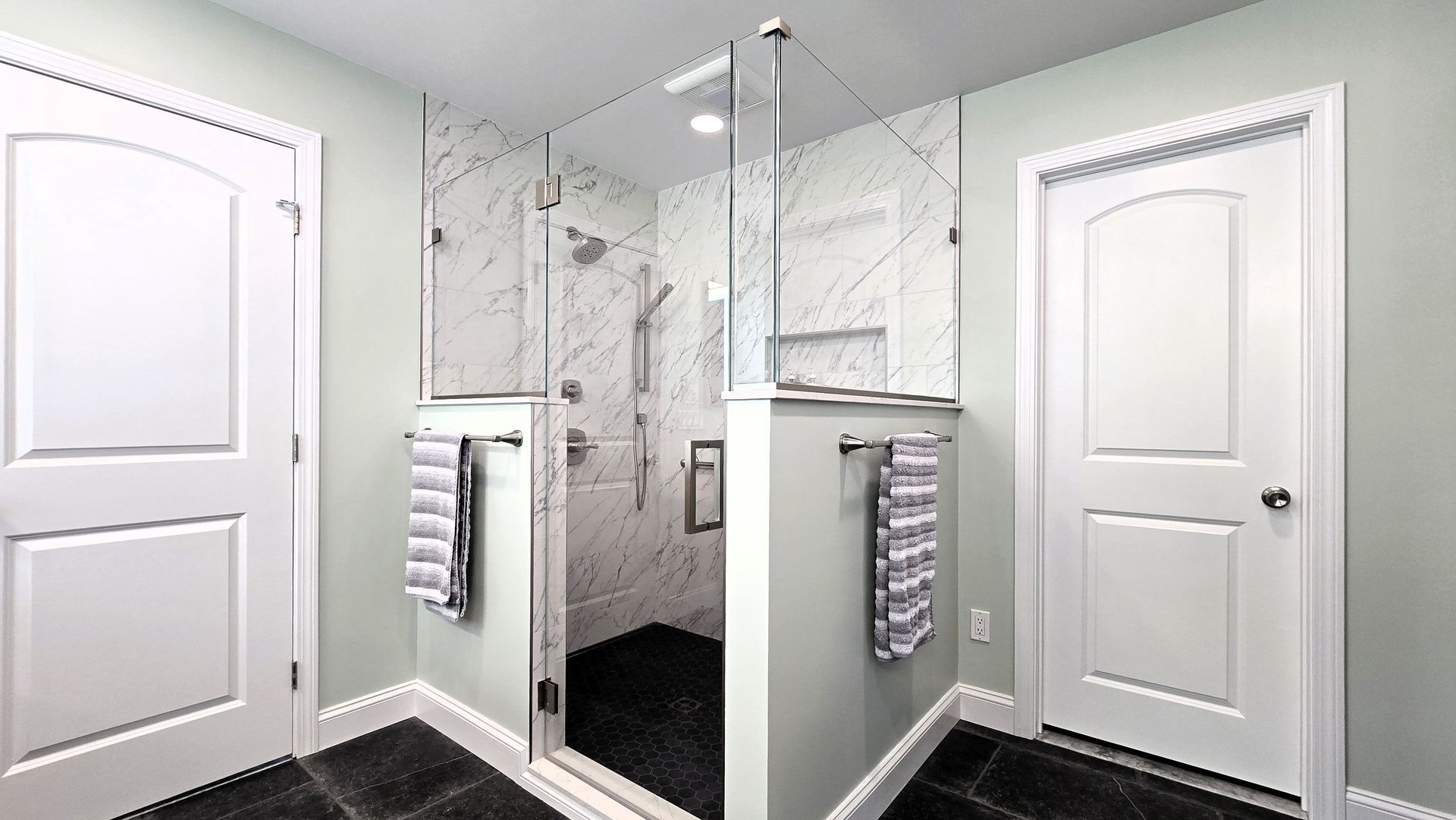 A modern bathroom in Glastonbury, CT featuring a white vanity with marble countertop, light green walls, black floor tiles, and a glass shower.