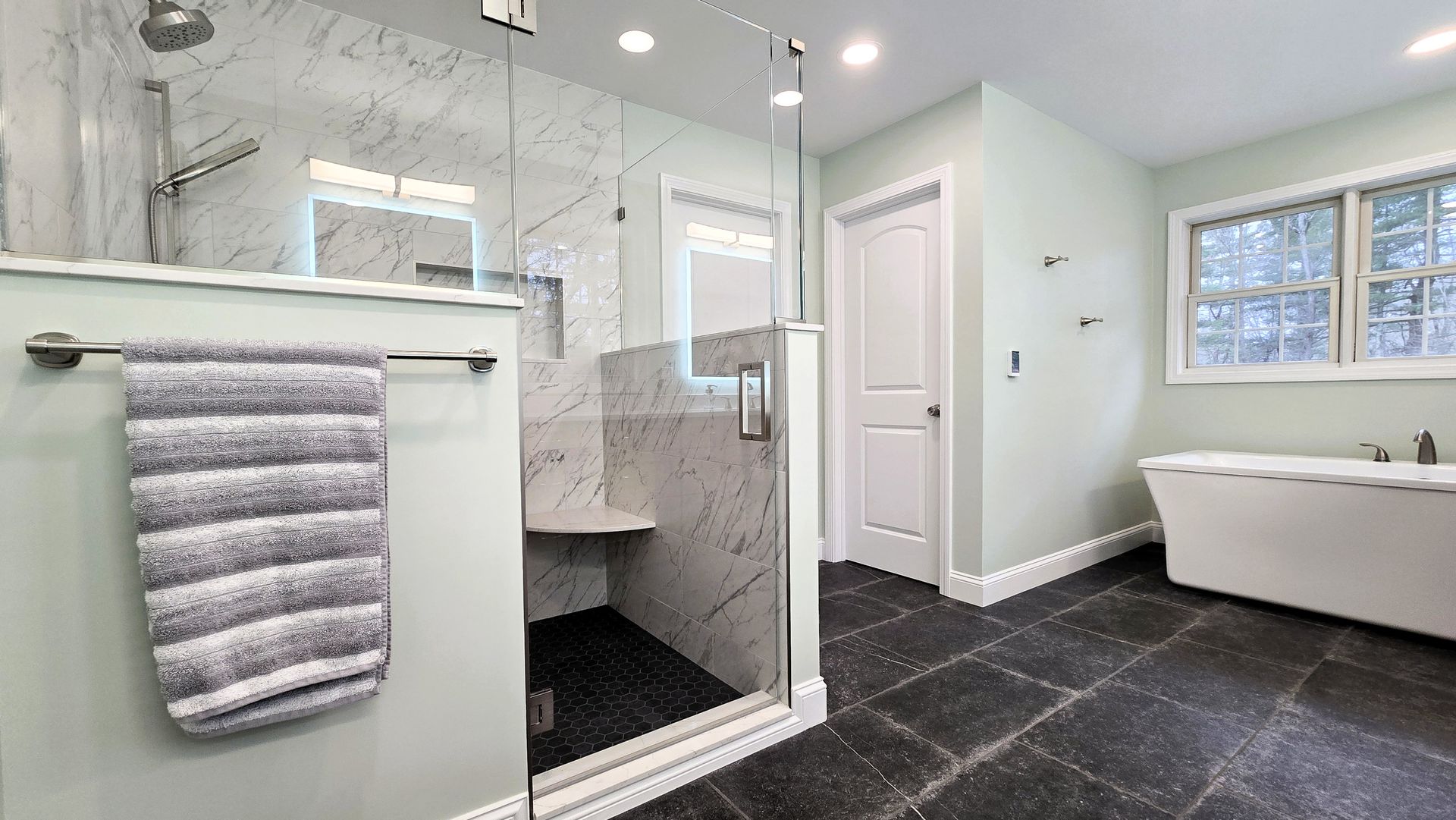 A modern bathroom with a glass-enclosed shower in Glastonbury, CT, a white soaking tub, and dark tiled floors.