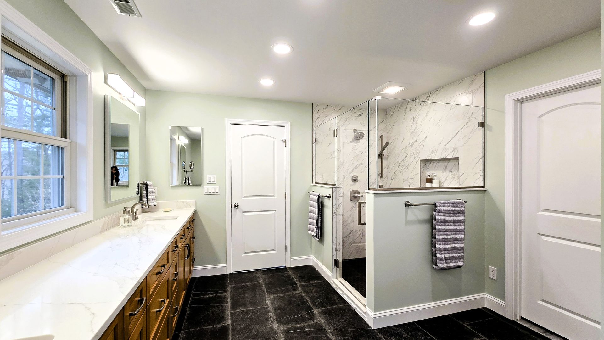 A vanity with a slatted wood finish and gold handles stands in a white bathroom in Cromwell, CT next to a window and a wall mirror.