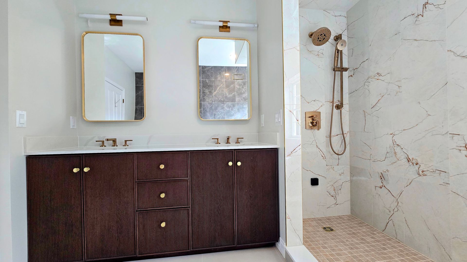 A vanity with a slatted wood finish and gold handles stands in a white bathroom in Cromwell, CT next to a window and a wall mirror.