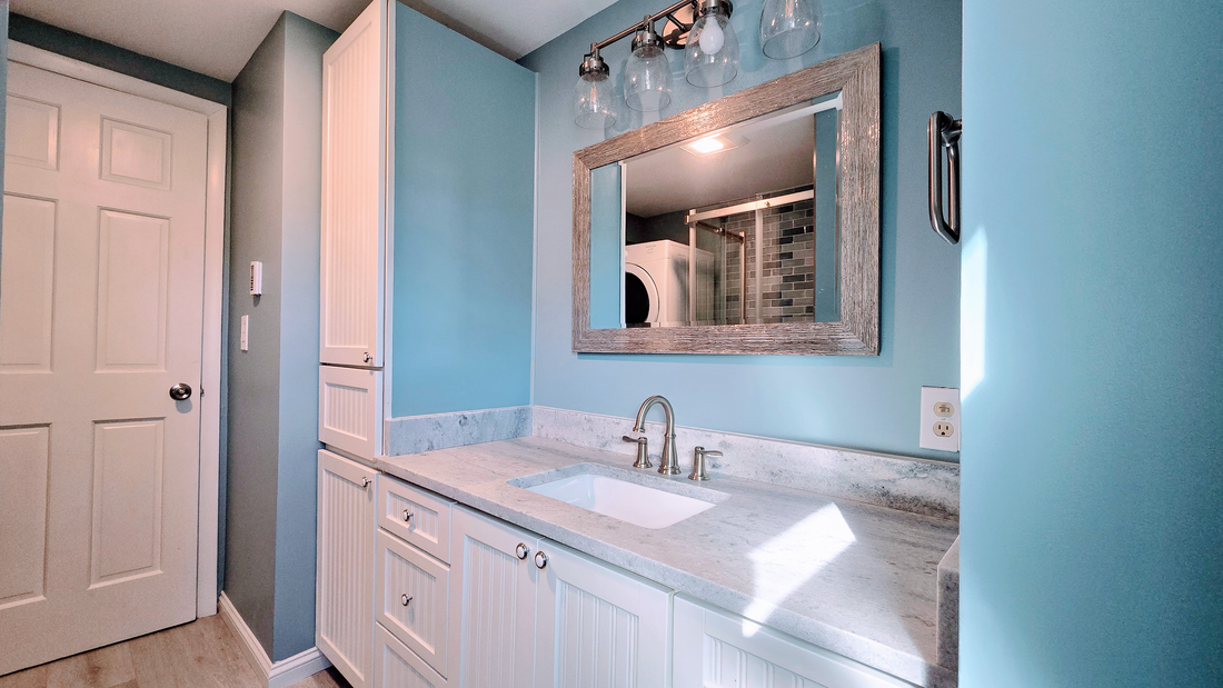 Bathroom remodel in Ashford, CT completed by First Choice Bathroom Remodeling with blue walls, white cabinets, marble countertop, large mirror, and a closed white door.