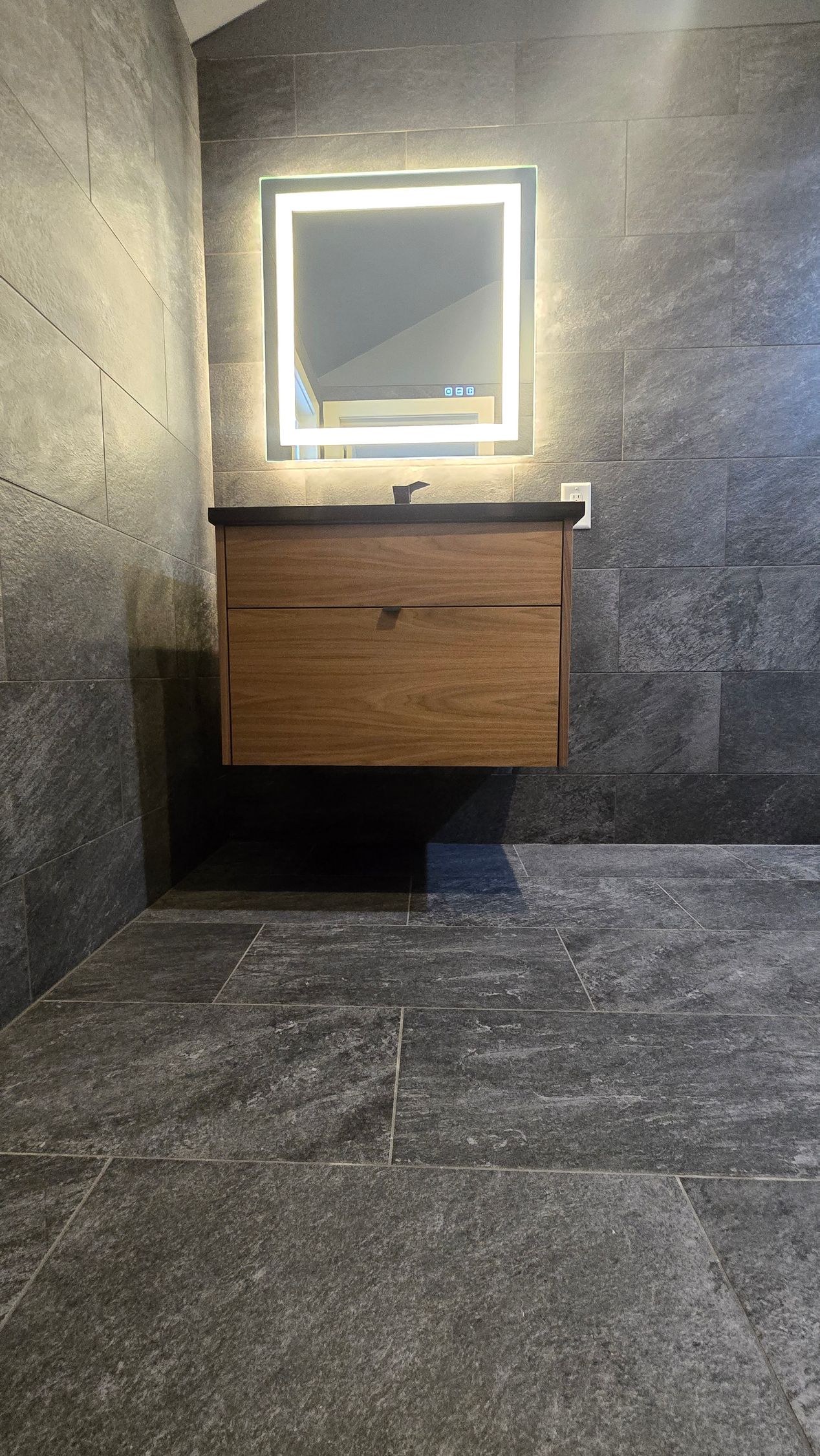 A floating wood-grain vanity under an illuminated square mirror against a dark, textured stone tile wall and floor.