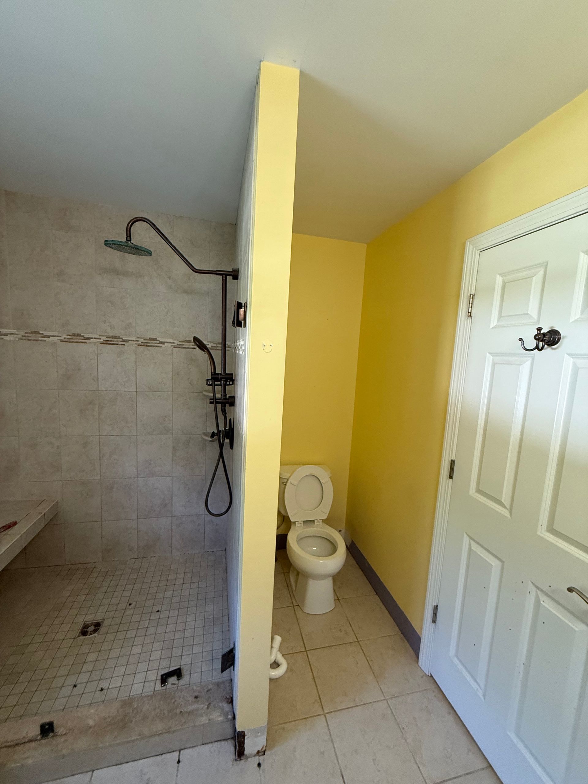 Bathroom with shower on left, toilet and door on right, divided by a yellow wall.