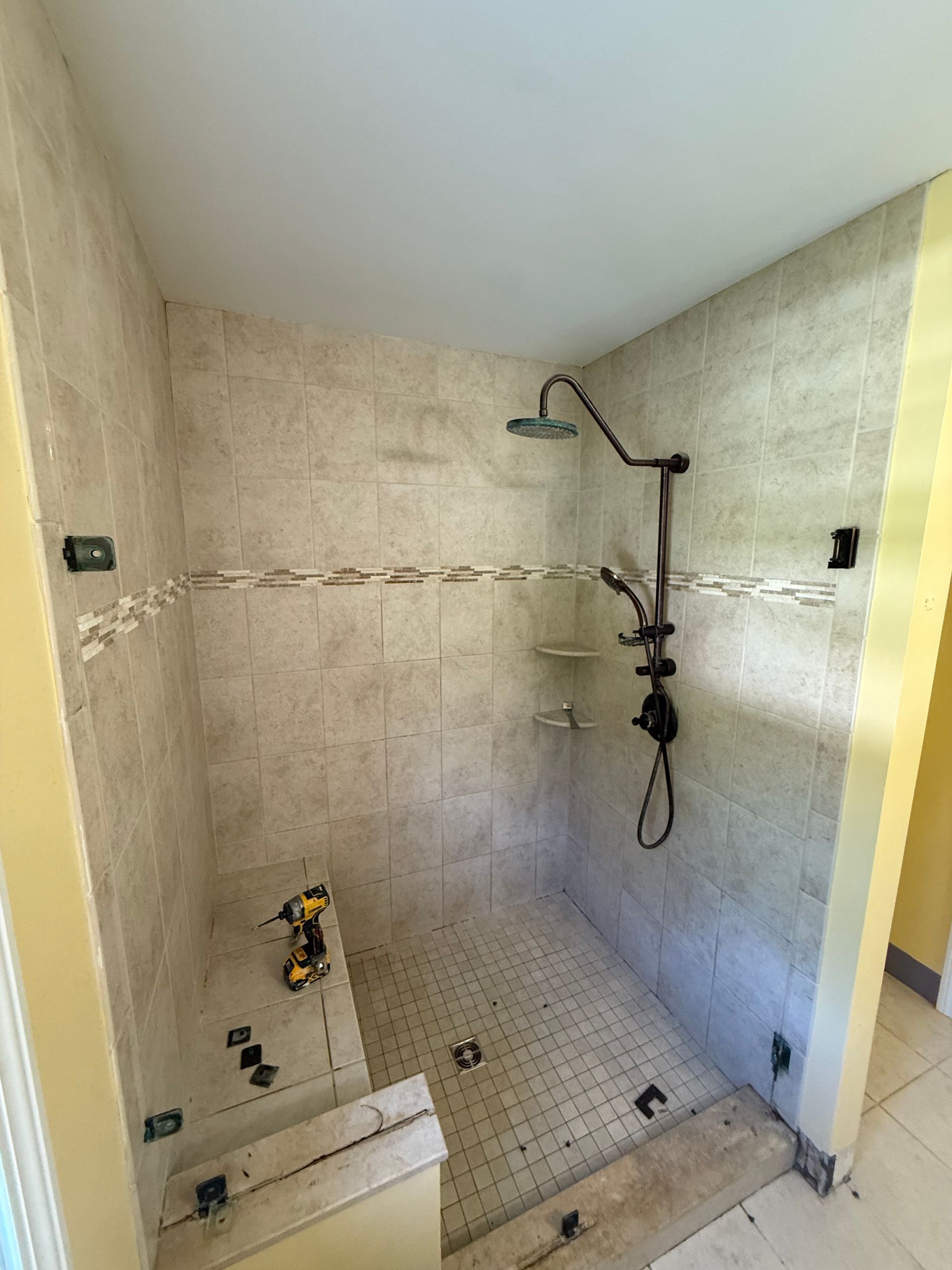 Shower stall with tan tile walls, dark shower fixtures, and a partially tiled floor.