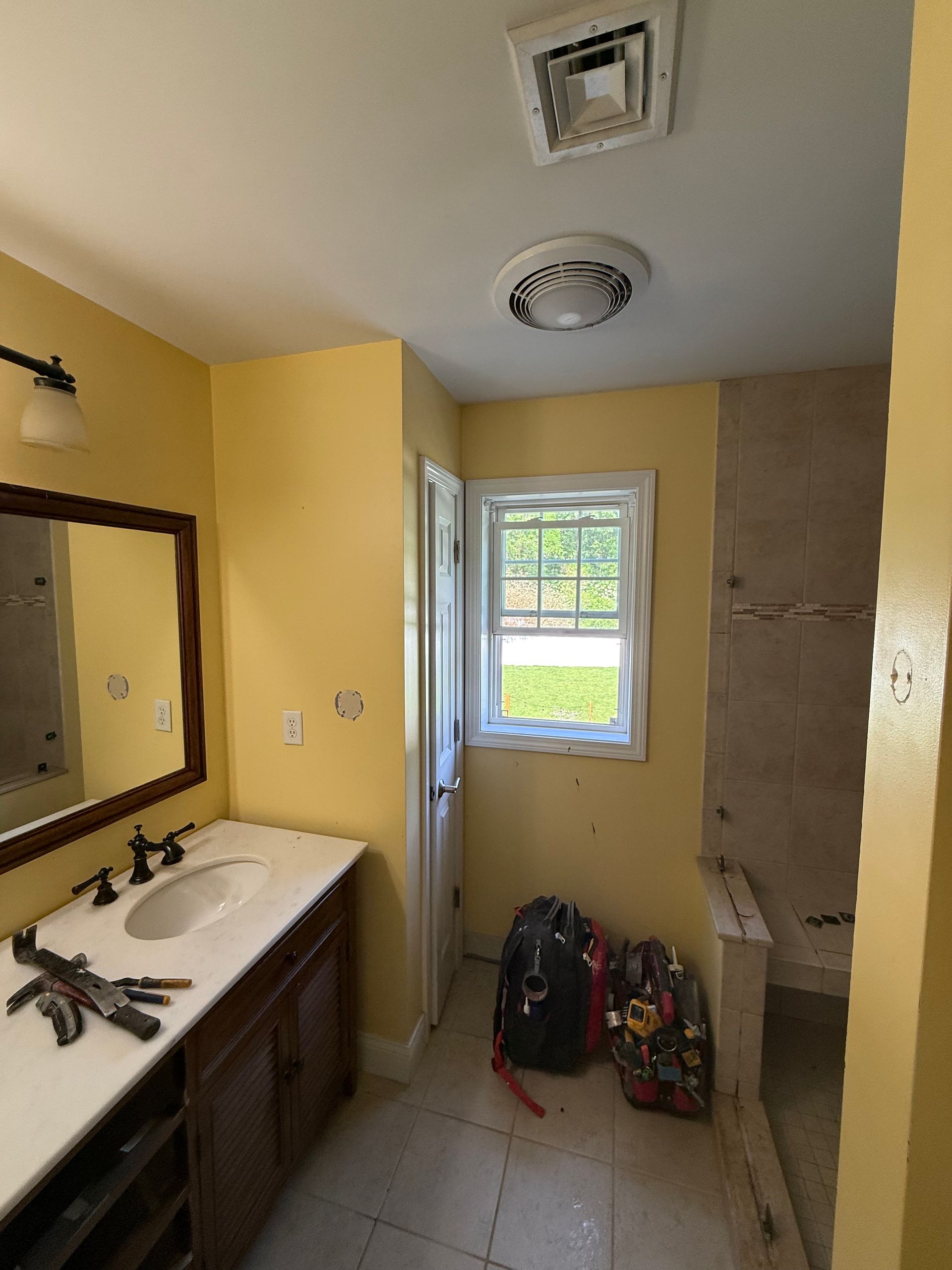Bathroom renovation in progress: yellow walls, vanity, window, open door, tile floor, bags on floor, ceiling vents.
