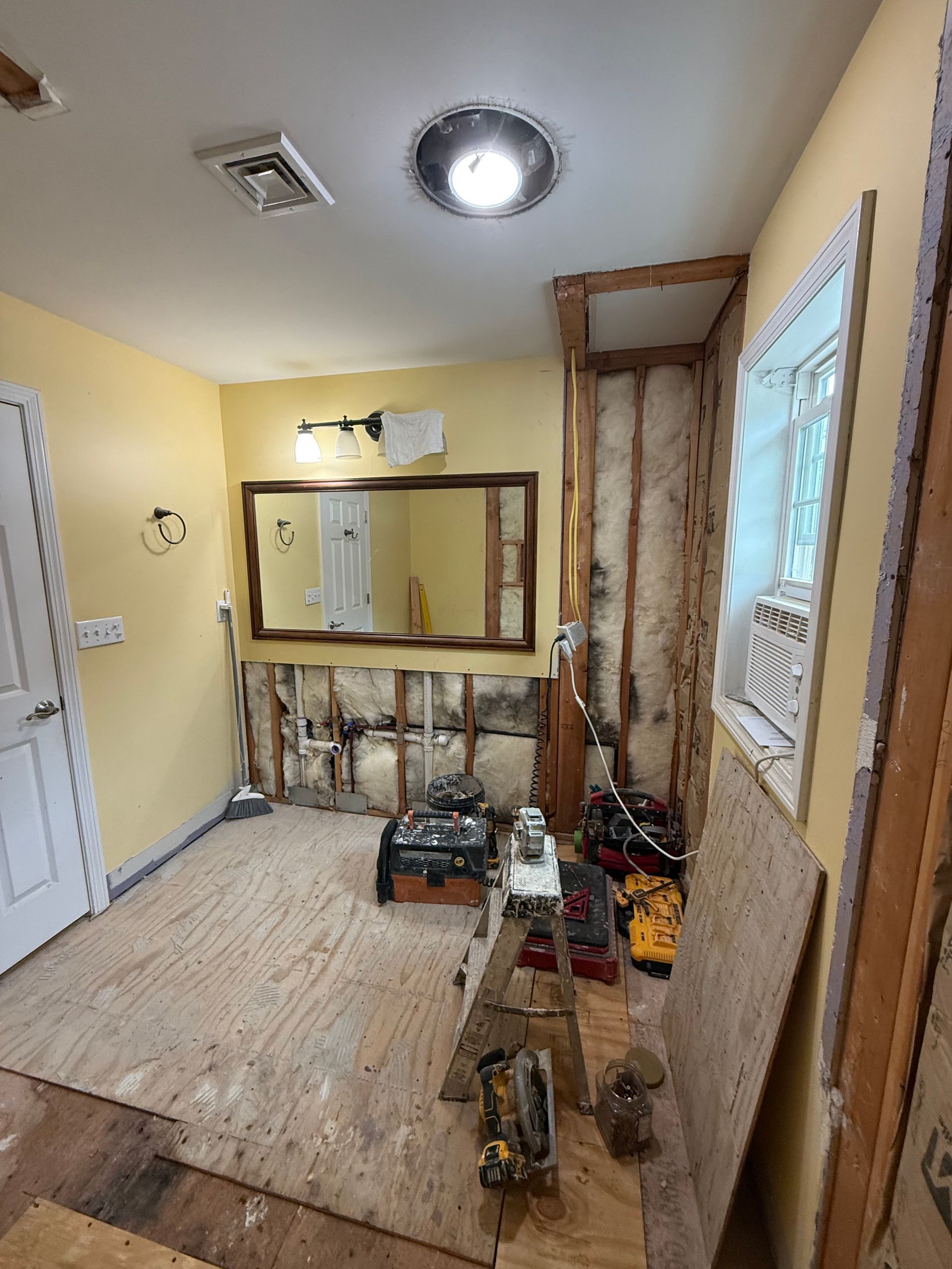 Bathroom under renovation; exposed studs, plywood flooring, tools, and large mirror.