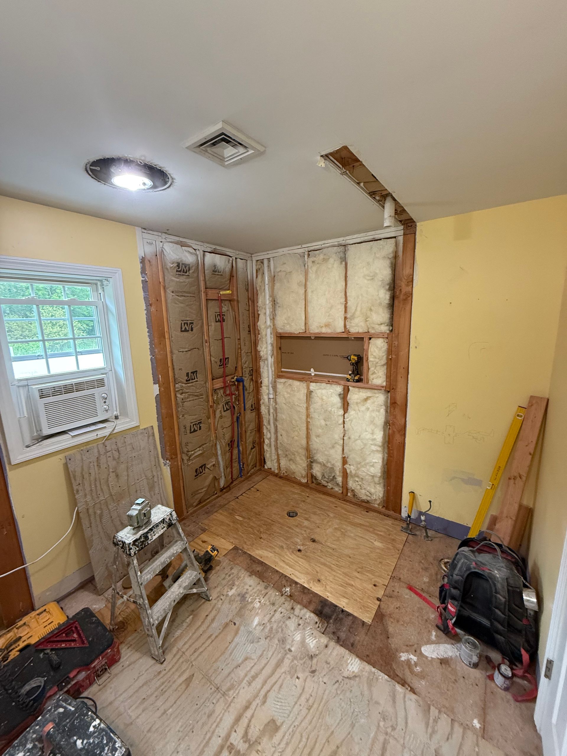 Bathroom renovation in progress; exposed studs, insulation, plywood floor, yellow walls.