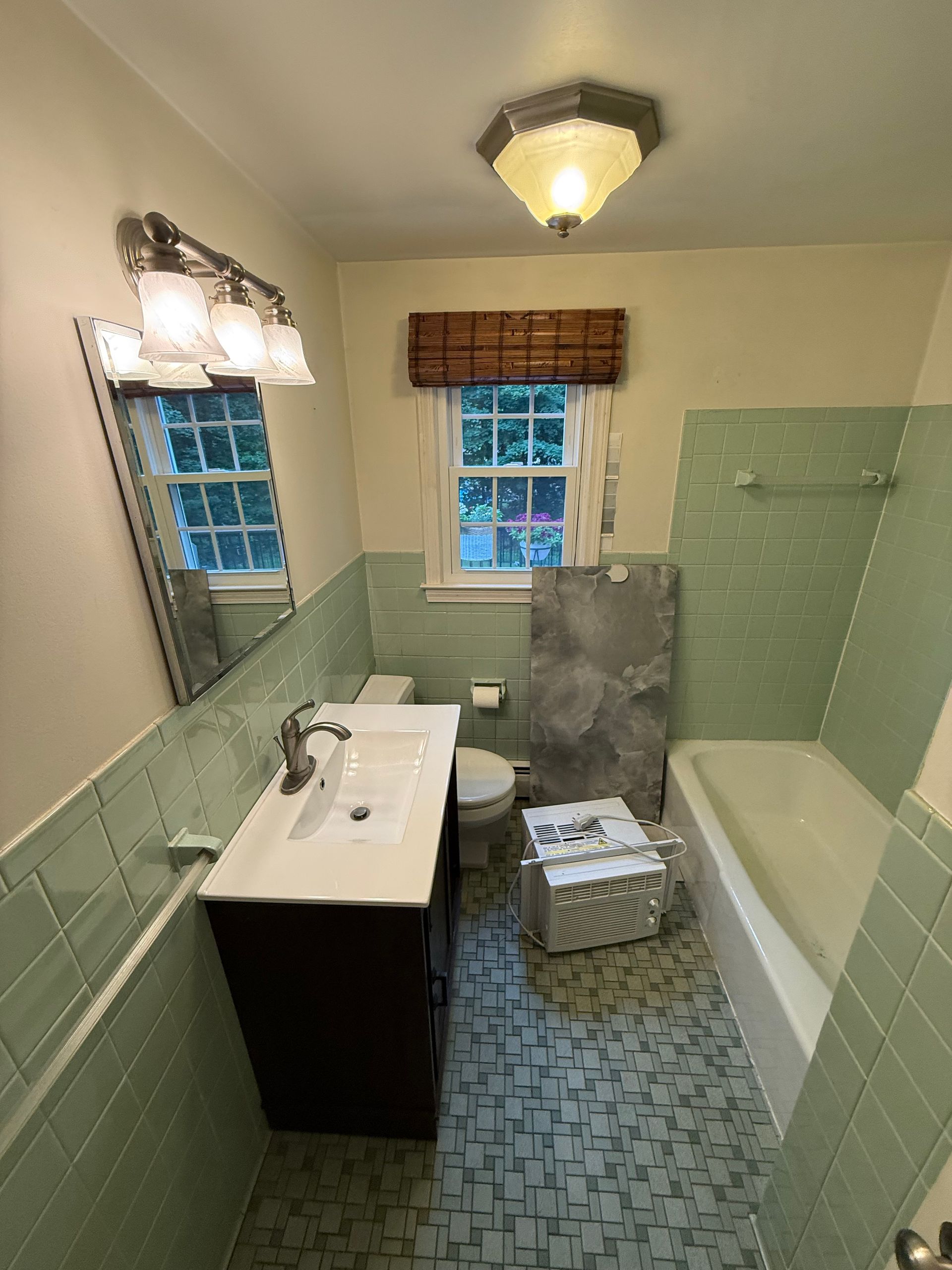 A bathroom with green tile, a dark vanity, and a window with a bamboo shade. A mirror hangs above the vanity with a three-light fixture.