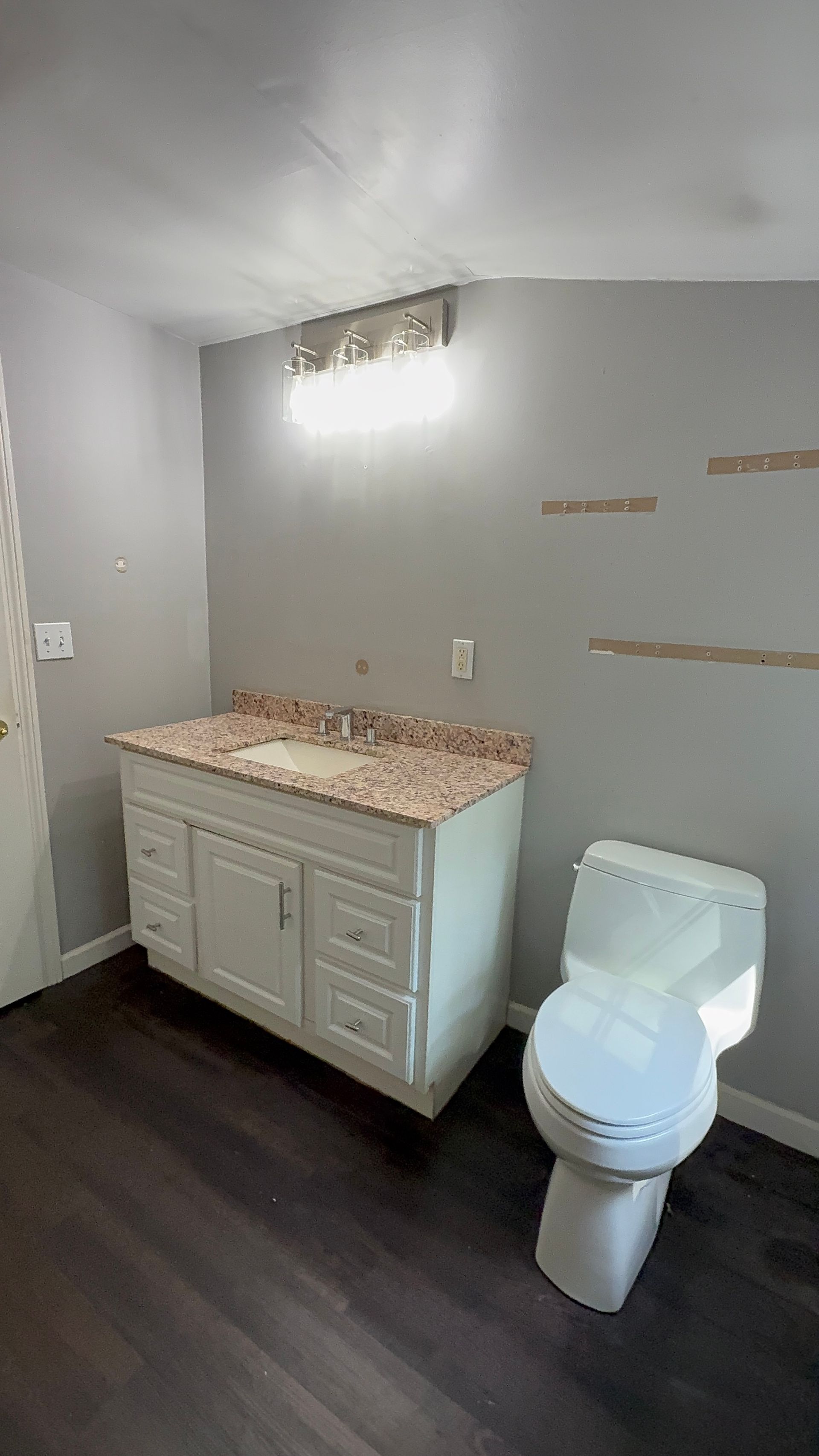 White bathroom with vanity, toilet, and dark wood-look floor. Gray walls, with a light fixture.