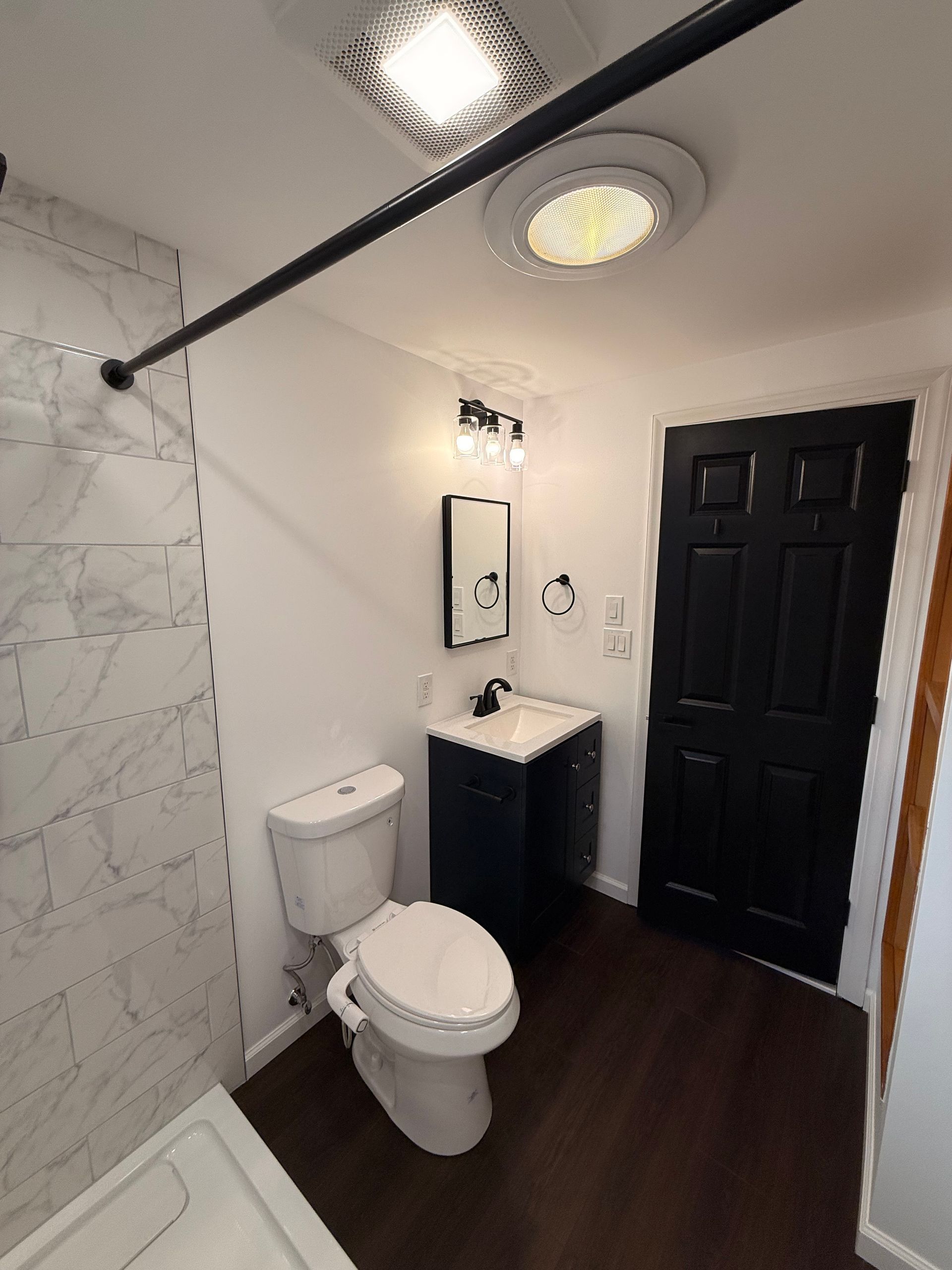 Small Berlin bathroom with marble-tiled shower, dark wood-look floor, black door and vanity, and white toilet.