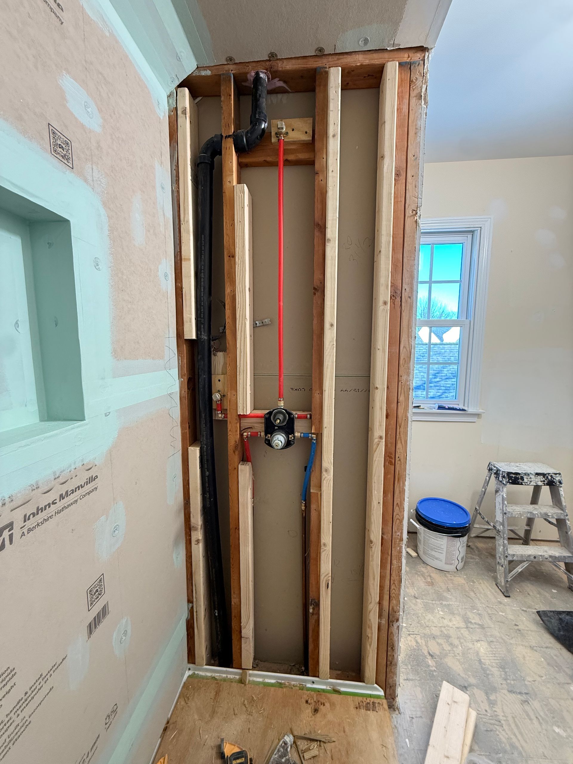 Exposed wall studs with plumbing pipes and a shower valve installed in a room under construction.