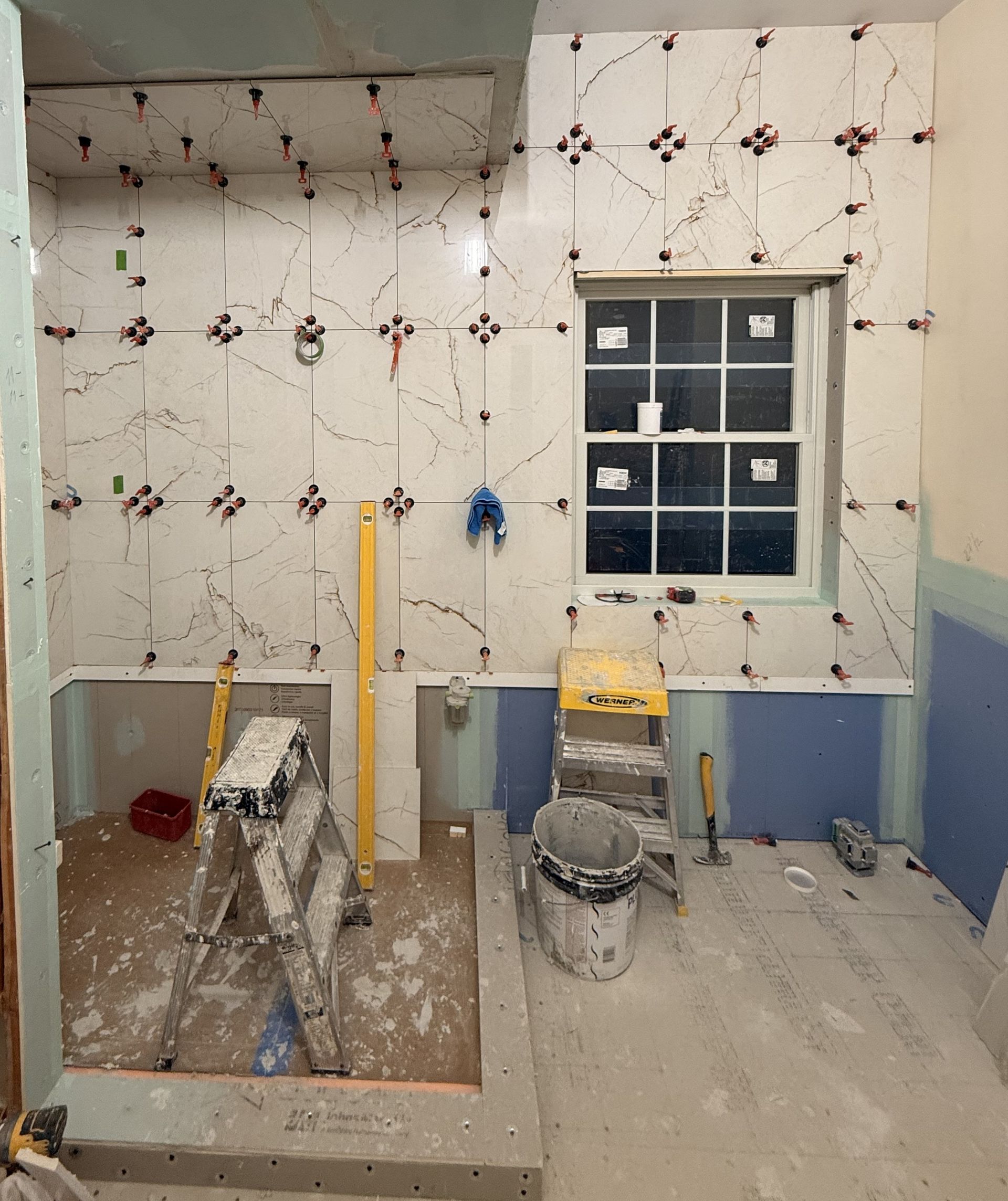 Construction site with unfinished tiled wall, window, and two step ladders in a dusty room.