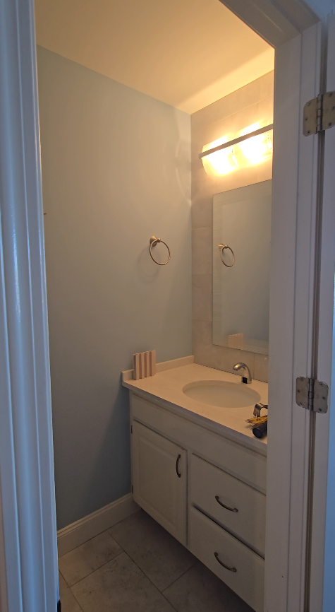 Small bathroom with a white vanity, sink, and mirror. Light blue walls, chrome towel ring, and a light fixture.
