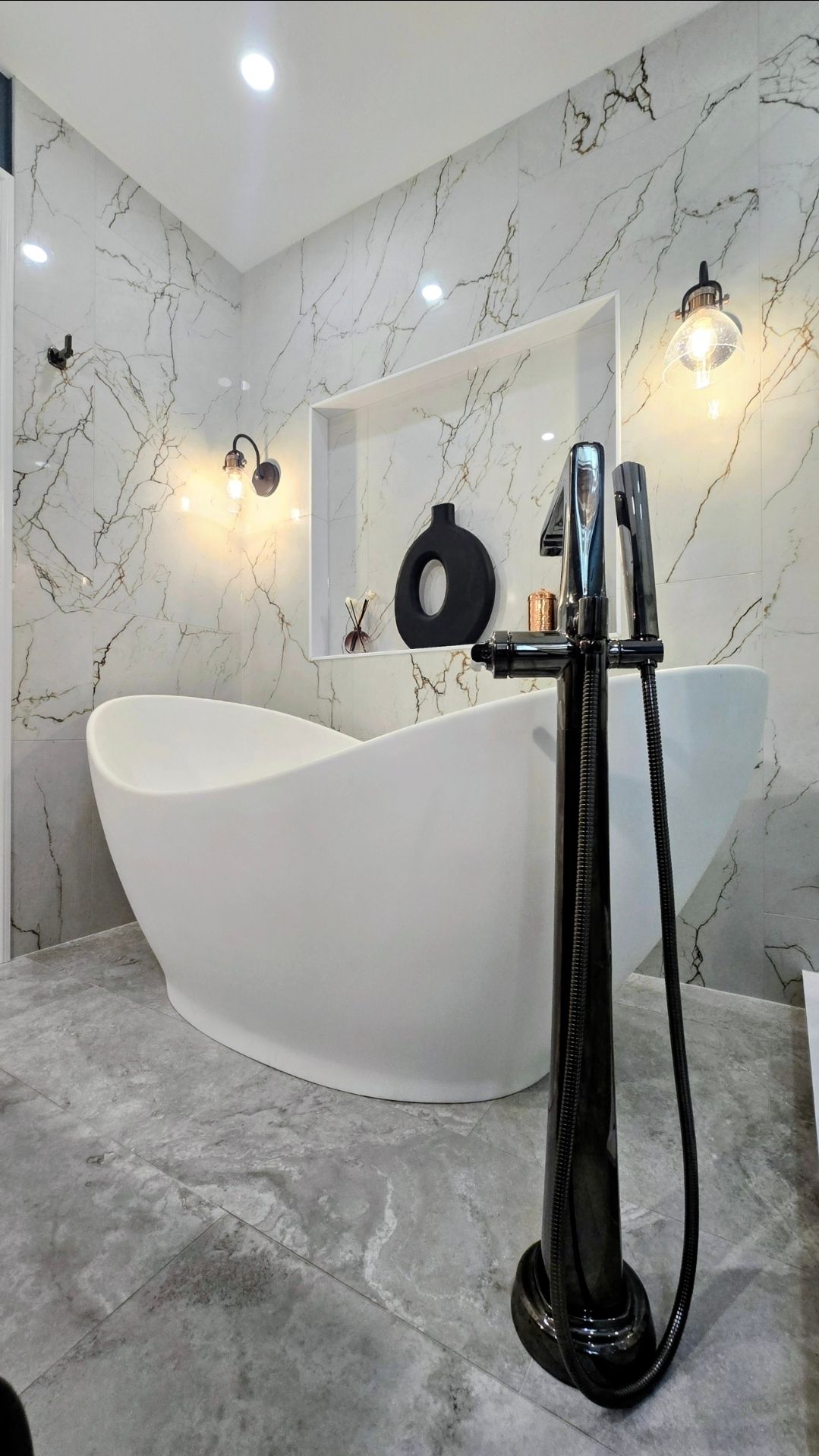 Modern bathroom renovation in Avon, CT with a white oval tub, black faucet, marble walls, and gray floor.