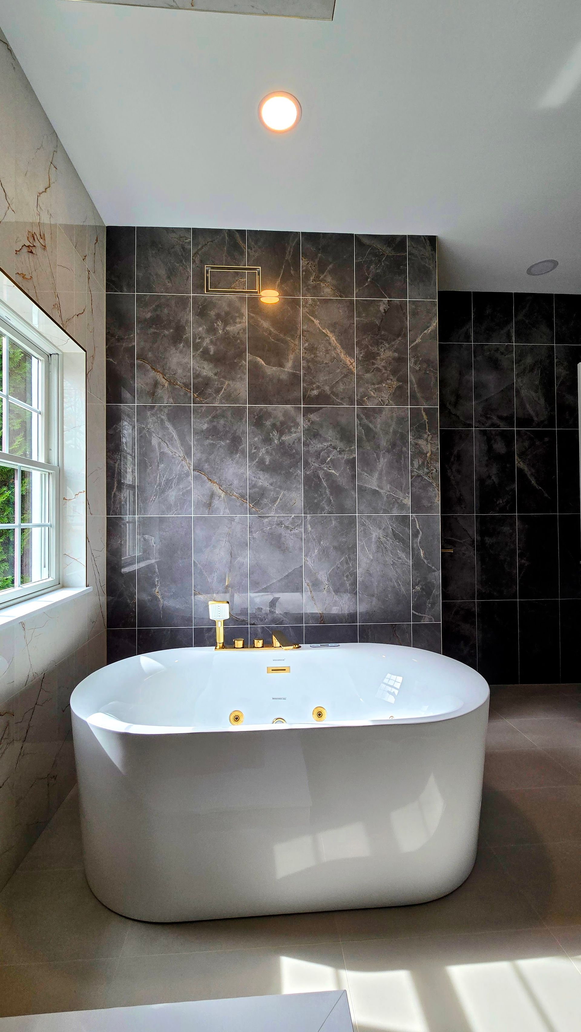 A modern white freestanding bathtub in Cromwell, CT in a bathroom with dark, metallic-inlaid marble walls and a side window.