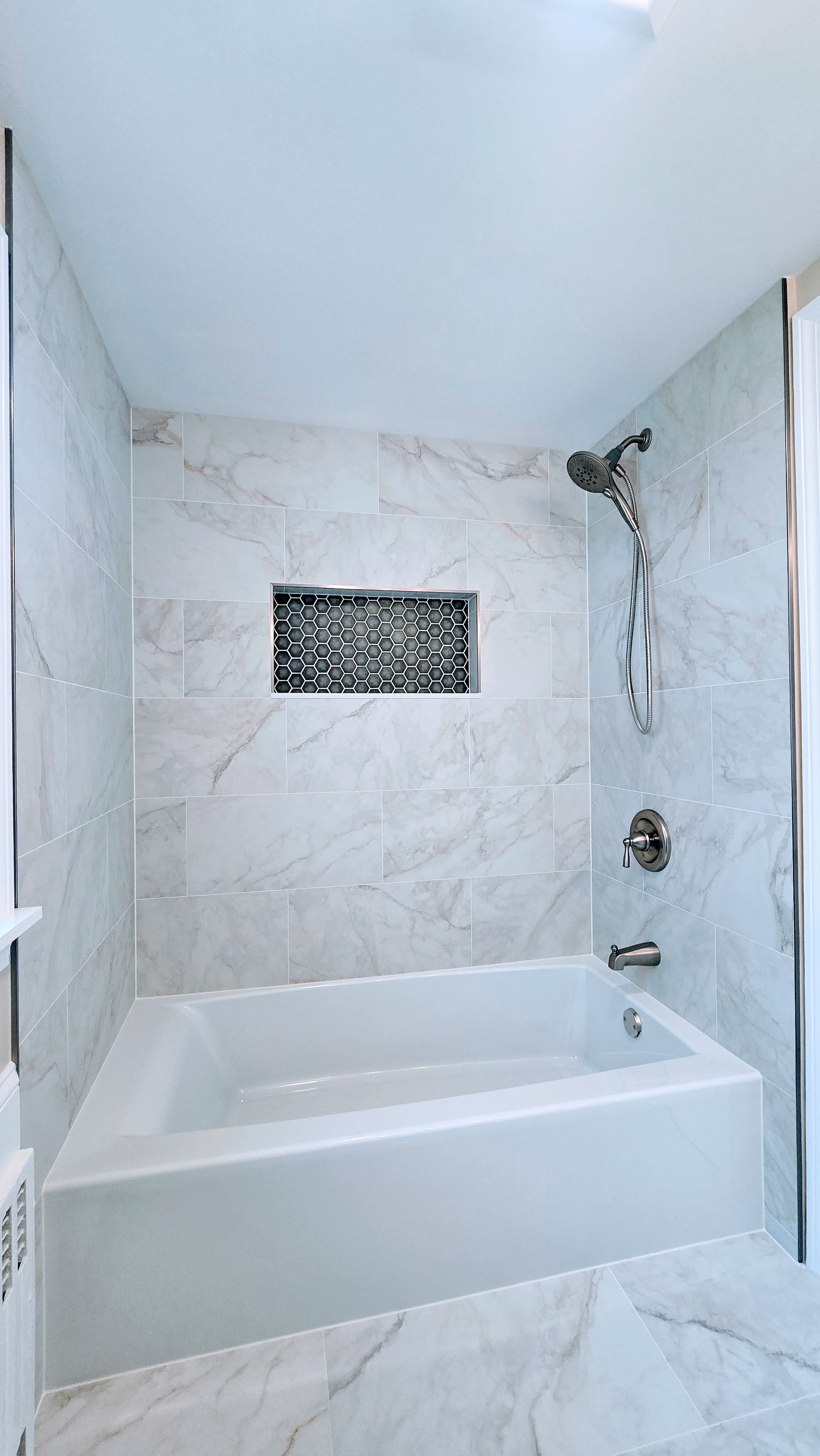Modern White tiled bathroom in Plantsville, CT completed by First Choice Bathroom Remodeling with a bathtub and showerhead. A dark accent in a wall niche.