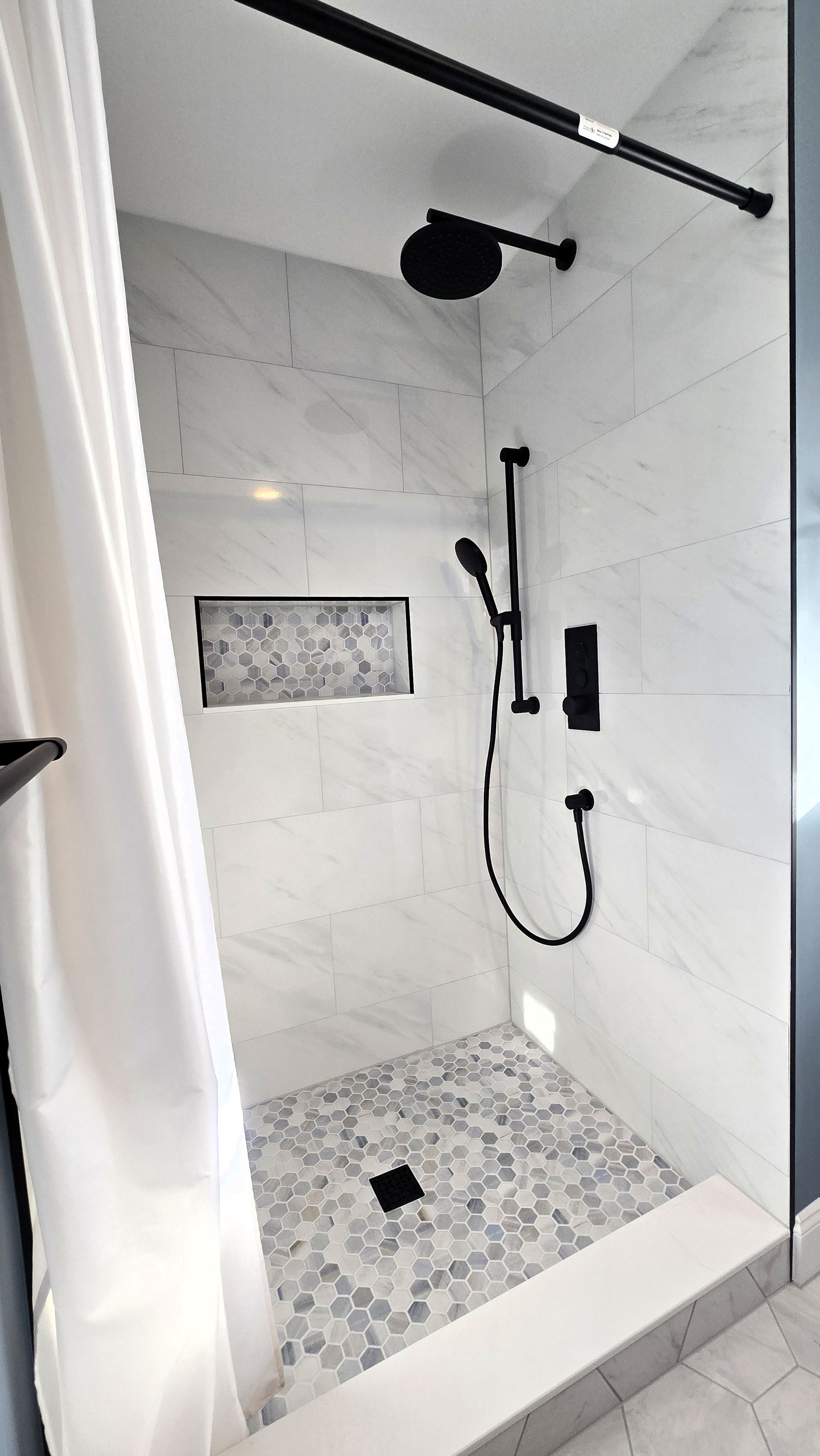 Modern shower in Newington, CT with white marble walls, mosaic tile floor, and black fixtures. A shower curtain hangs to the left.