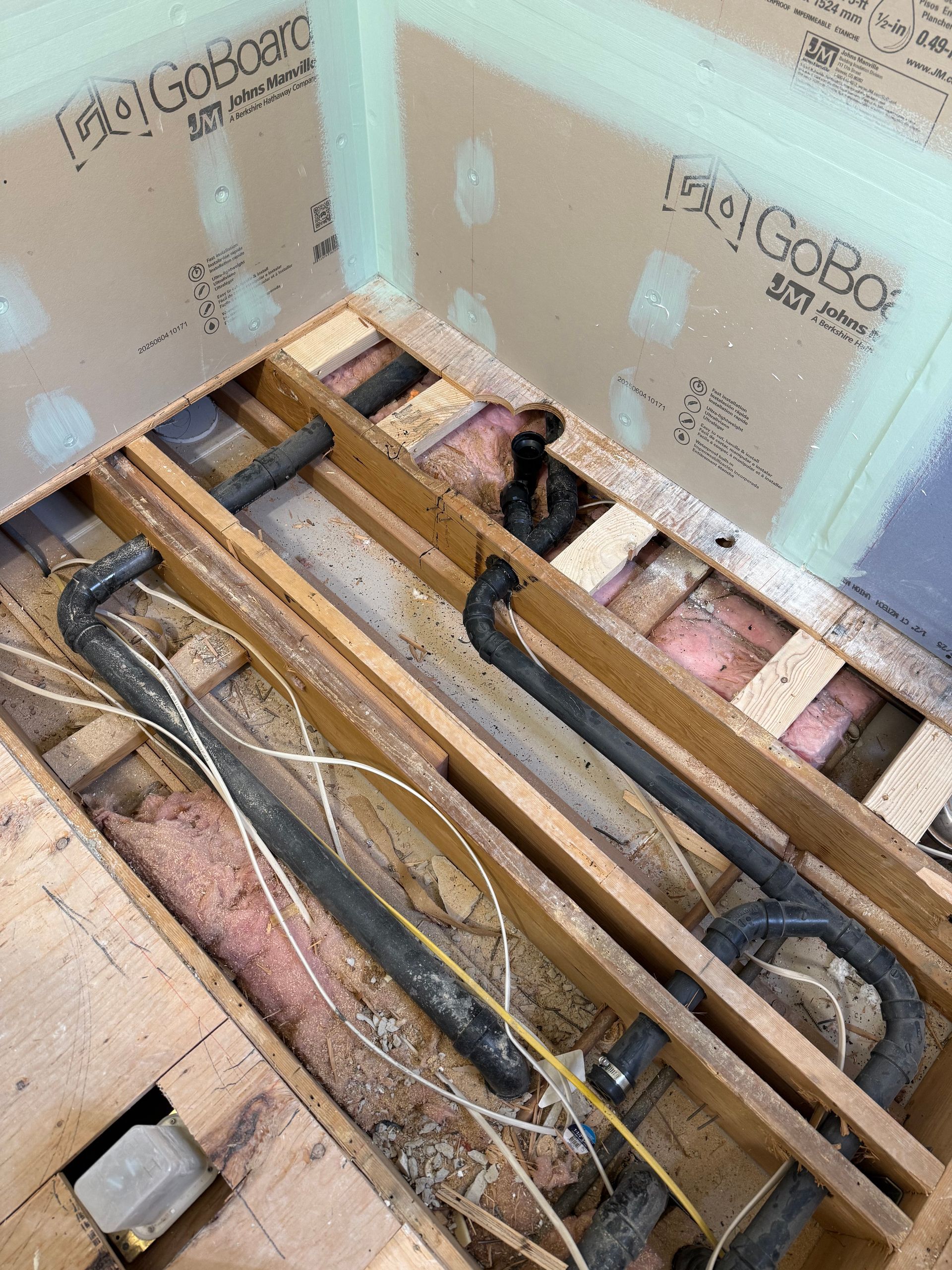 Framed floor joists with black pipes and wiring, ready for shower installation, with green GoBoard wall paneling.