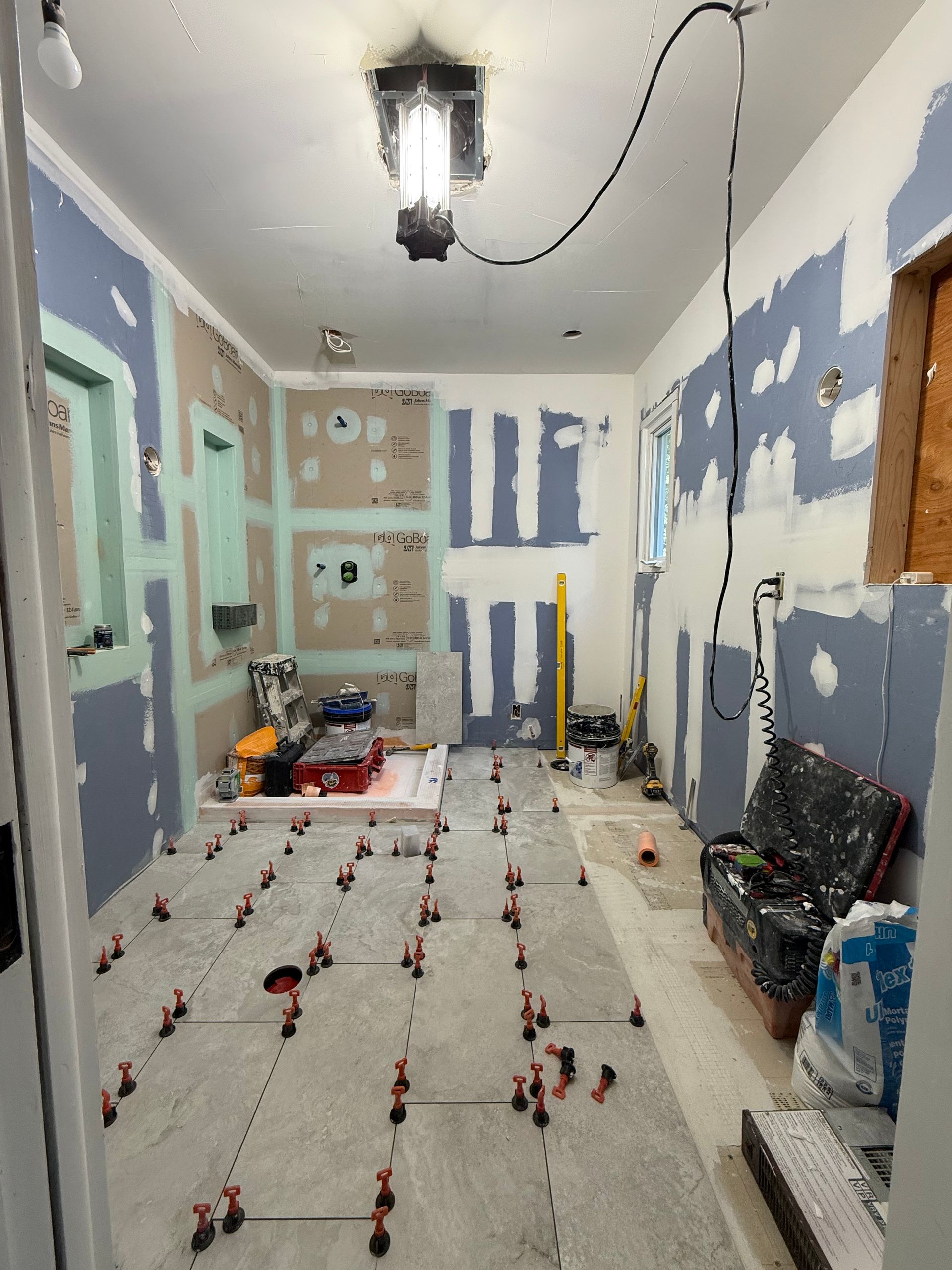 Bathroom renovation: tile floor in progress, drywall, tools, and temporary lighting.
