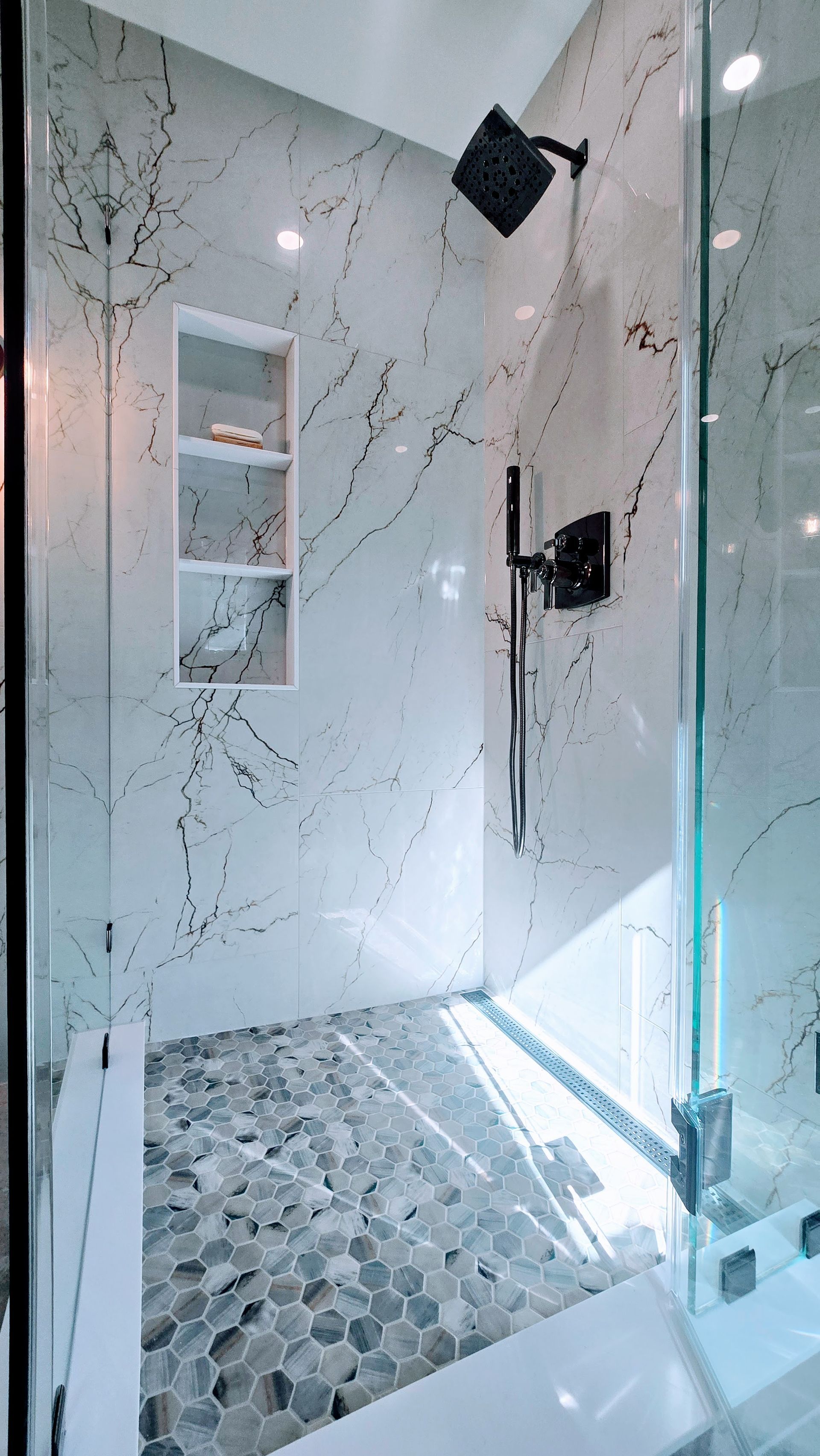 A modern walk-shower in Avon, CT with white marble walls featuring black veining, a pebble floor, a built-in shelf, and matte black fixtures.