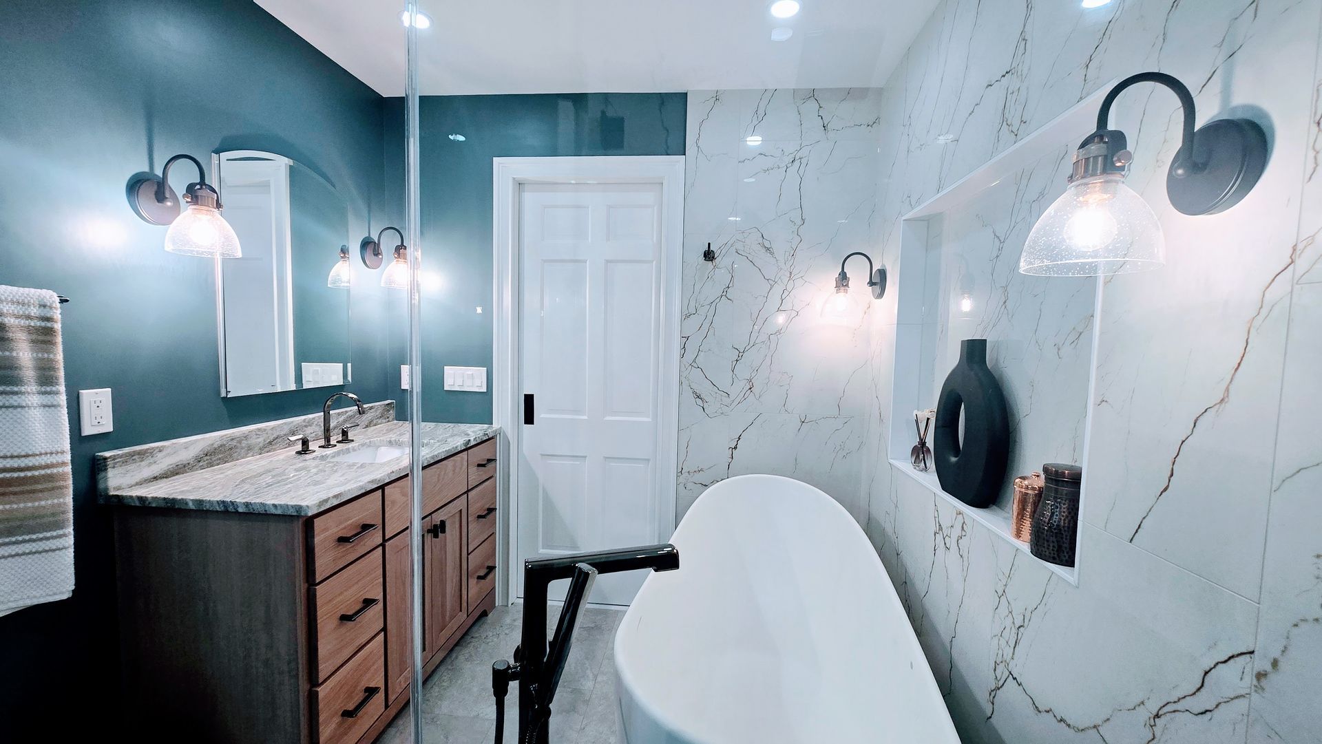 A modern bathroom remodel in Avon, CT featuring a white soaking tub, marble-tiled wall, vanity with wood cabinets, and dark teal accent walls.