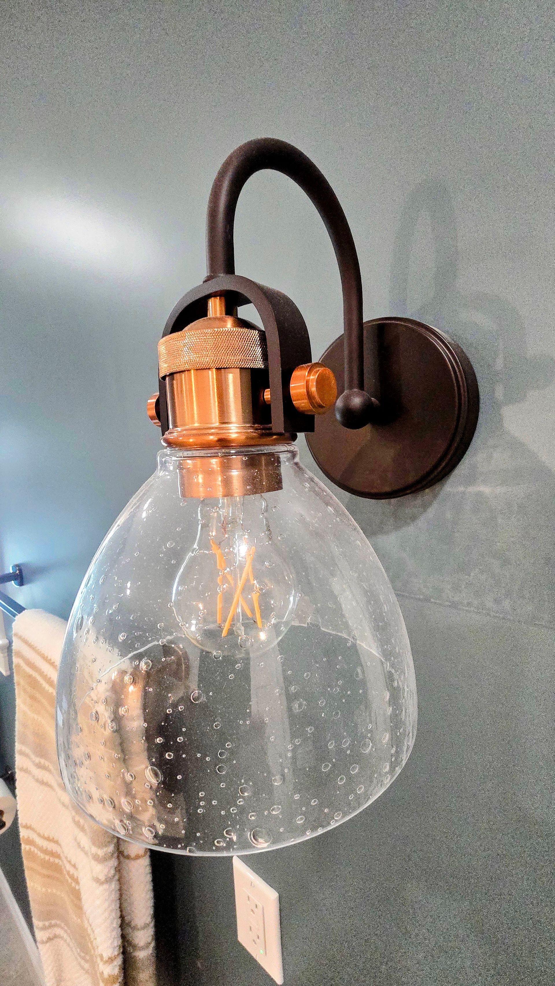 A seeded glass sconce with a copper bulb socket and dark metal hardware, mounted on a textured, dark gray wall.