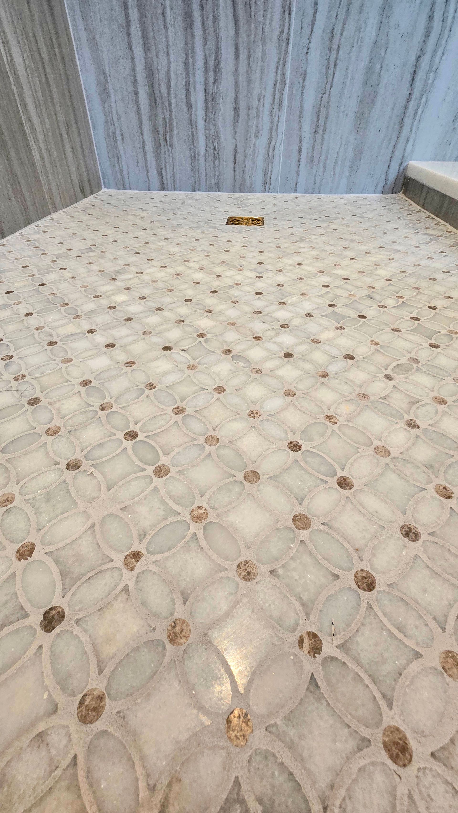 A tiled shower floor in Cheshire, CT with a white and cream floral-patterned marble design and a linear bronze drain.