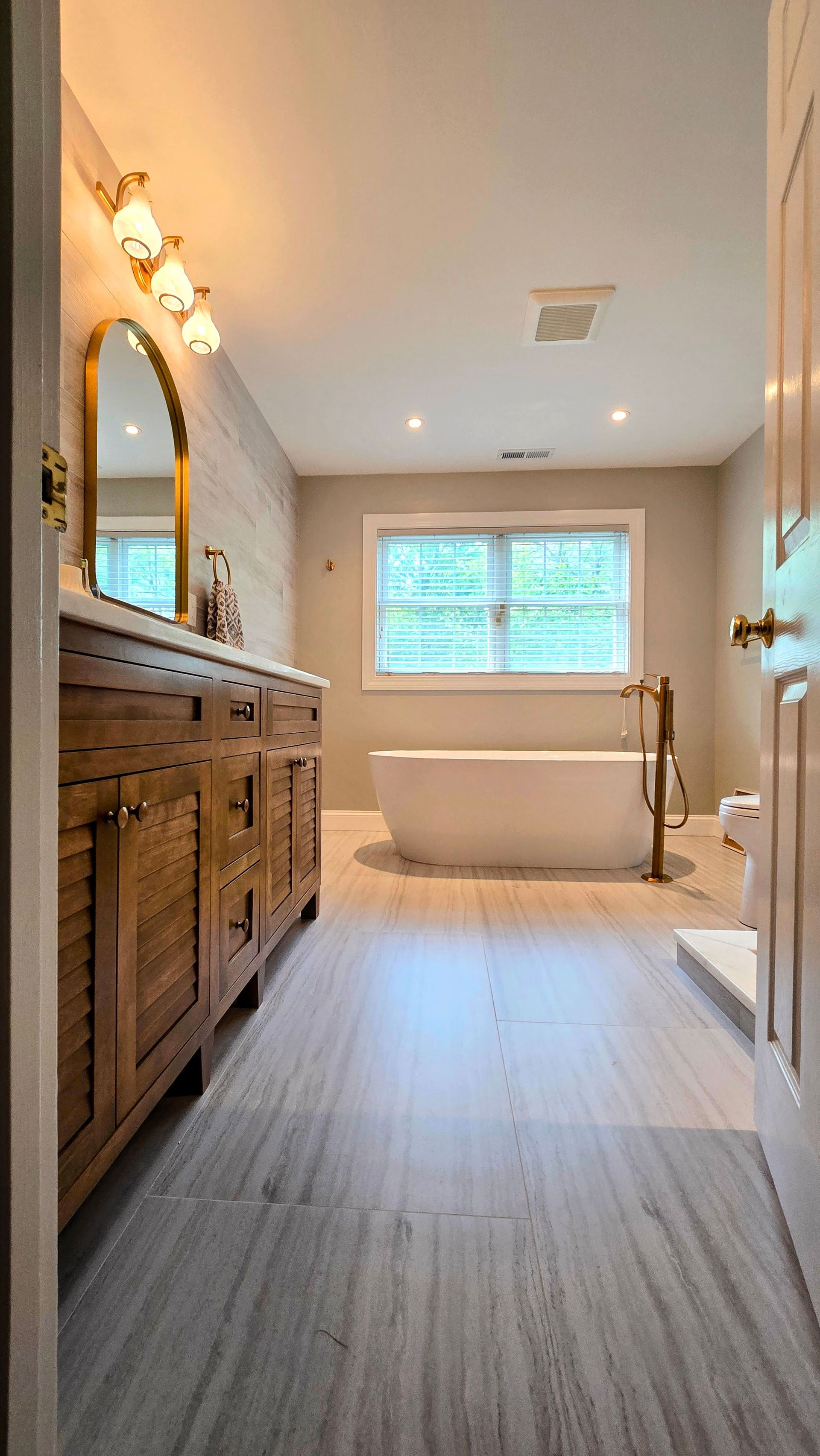 A bright, modern bathroom in Cheshire, CT with a wooden vanity, gold light fixtures, a freestanding tub, and light-toned wood flooring.