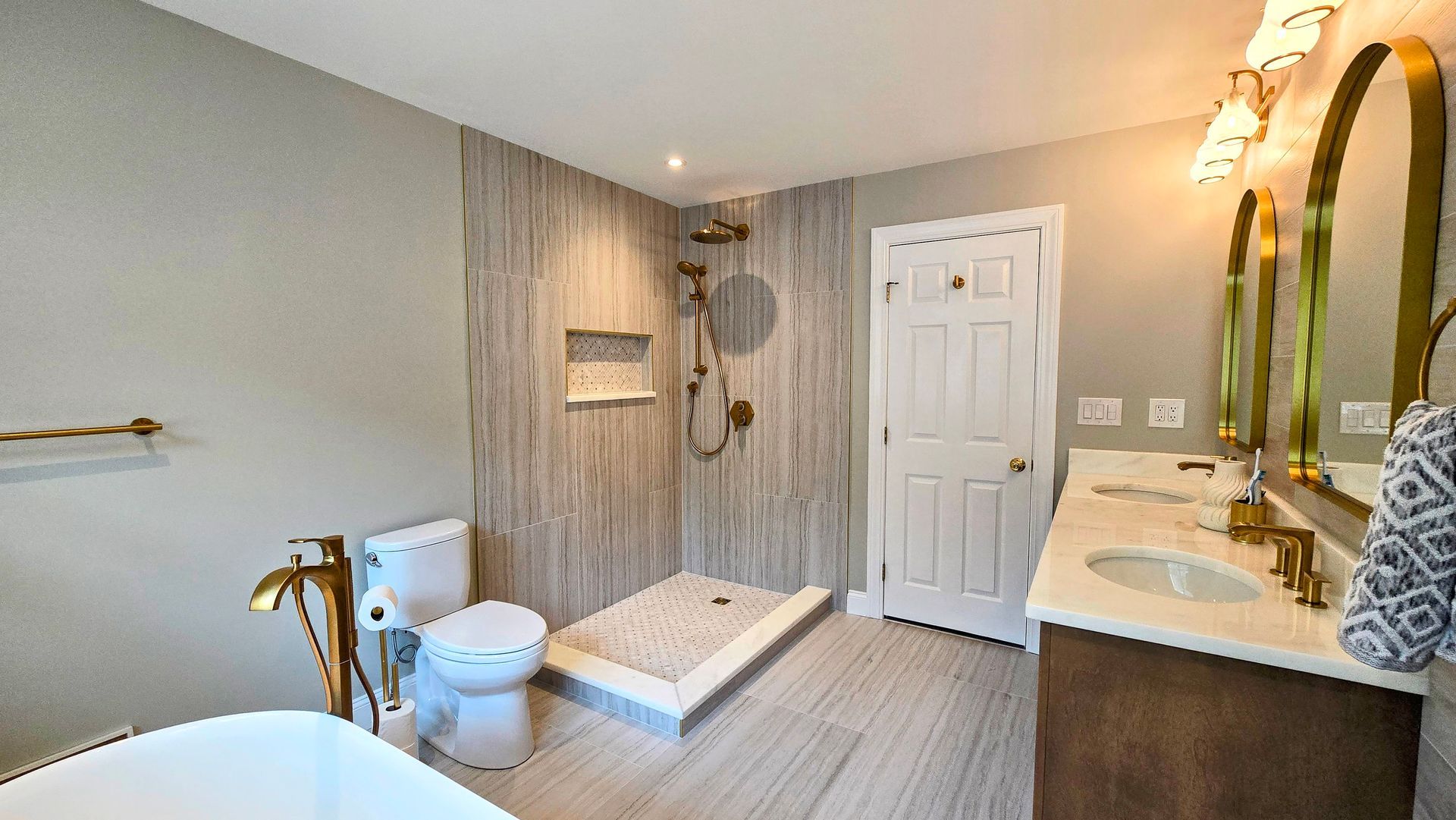 Modern bathroom with a freestanding tub in Cheshire, CT, toilet, walk-in shower with textured tile, and a double vanity with gold mirrors.