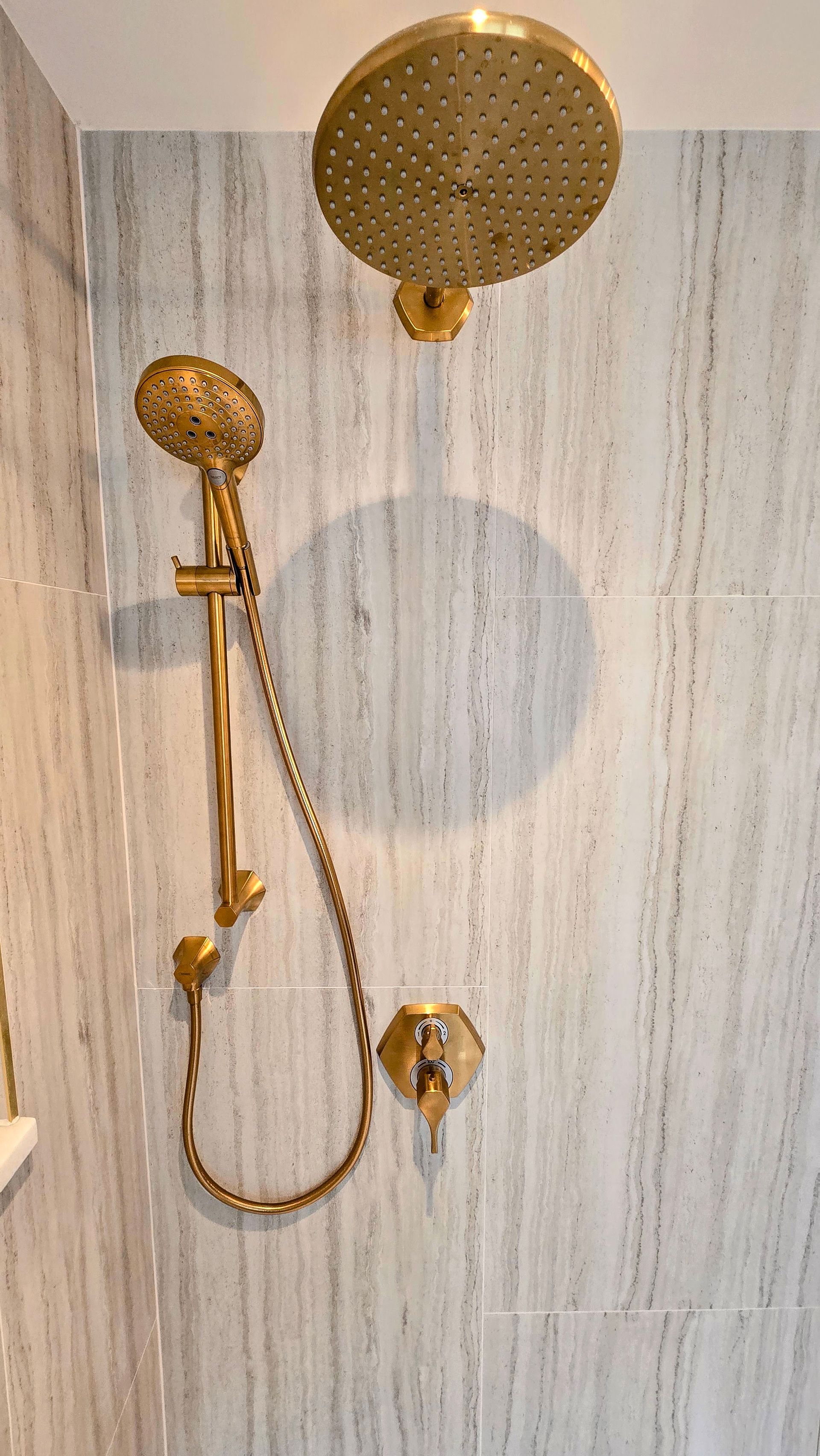 Gold-tone shower fixture with a large rain shower head, handheld sprayer, and wall-mounted control against marble tile.