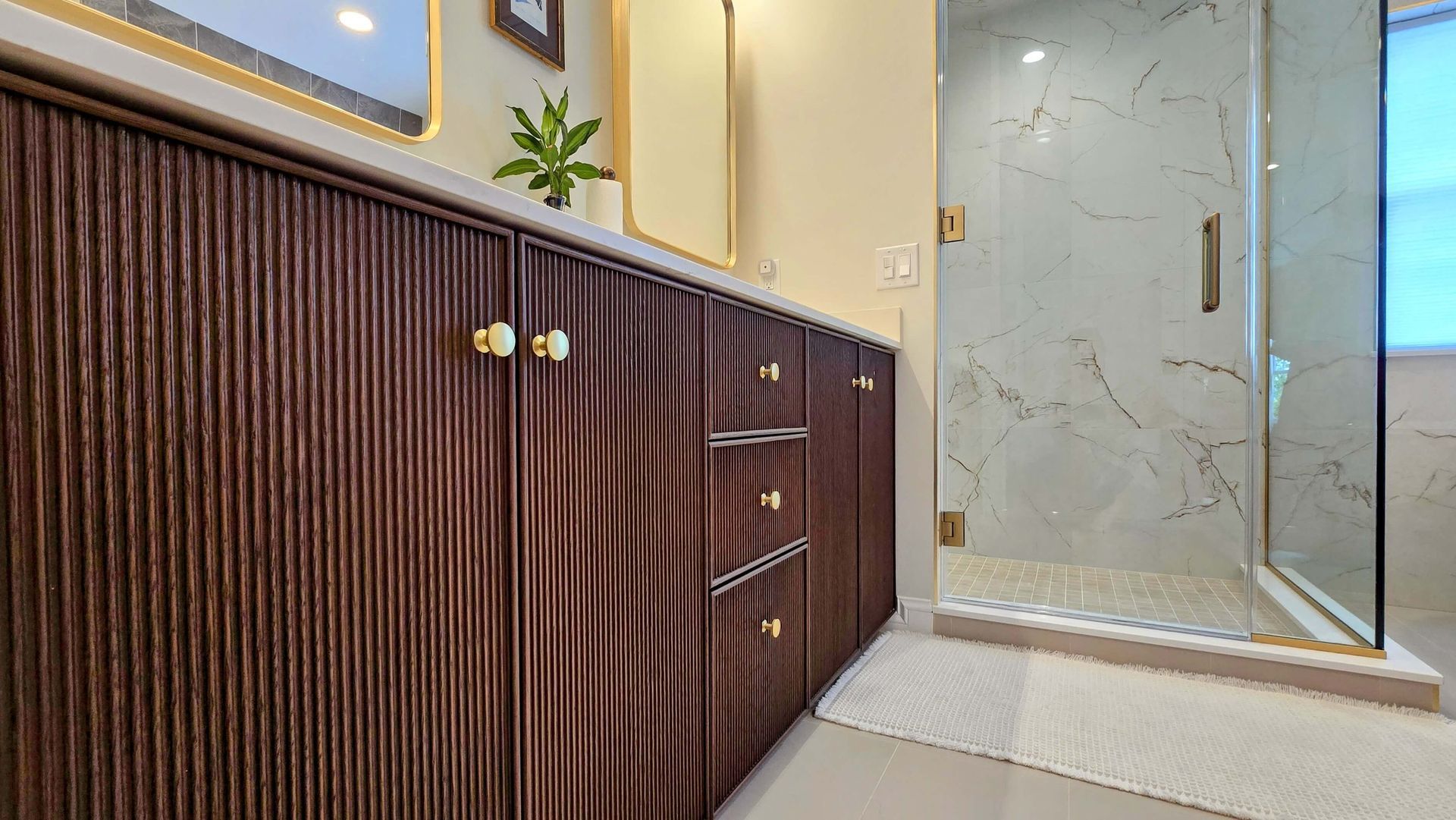 Modern bathroom vanity with dark wood cabinets, gold handles, and a glass shower door with marble tile flooring