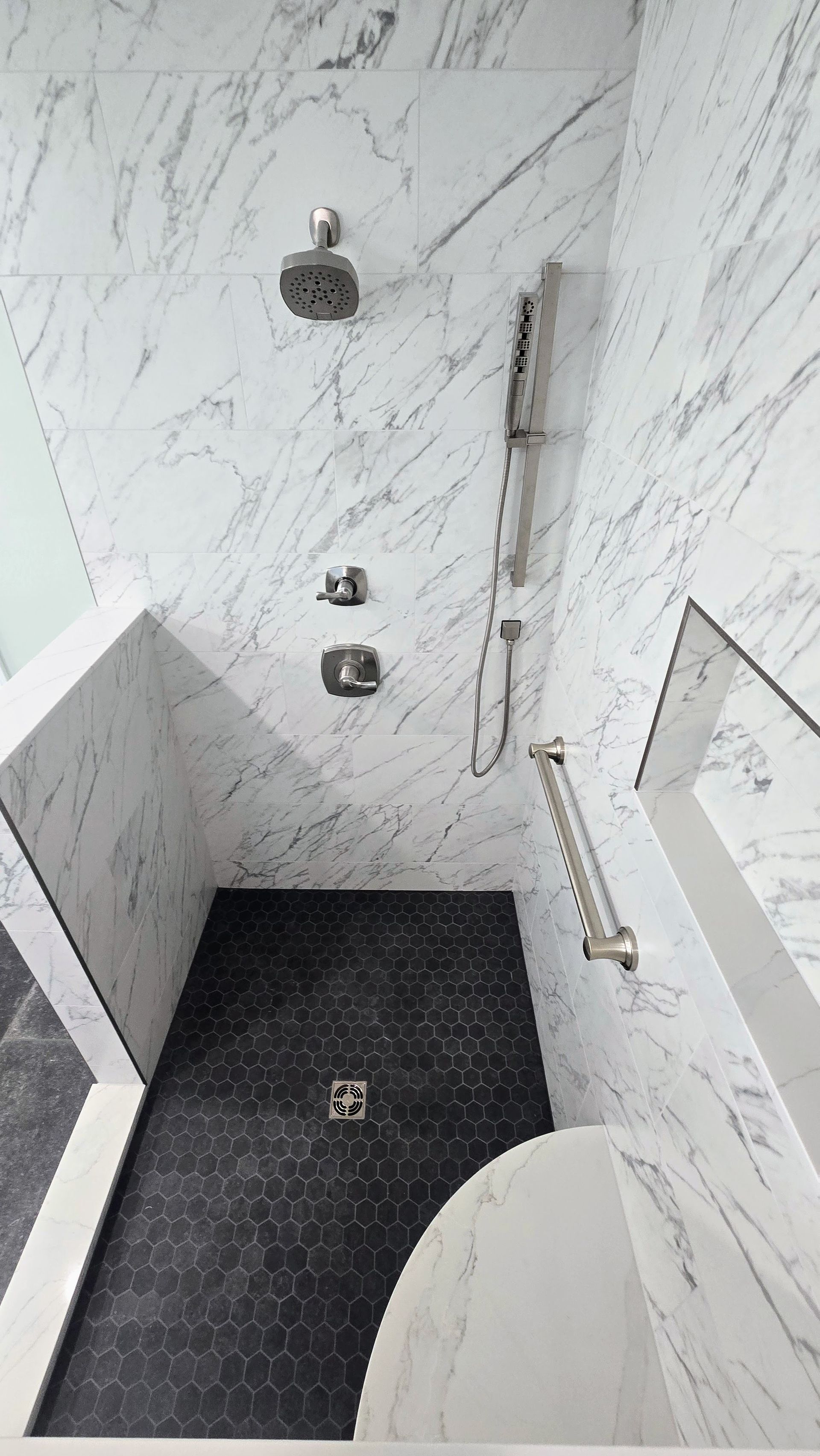 Modern walk-in shower in Glastonbury, CT with white marble-patterned tile walls, a dark stone floor, a chrome handheld sprayer, and grab bar.