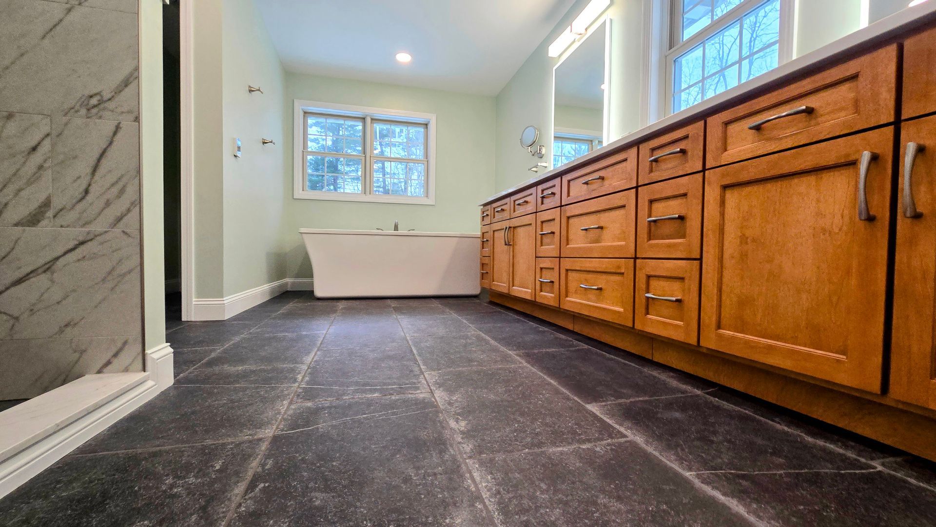 A modern bathroom in Glastonbury featuring a long wooden vanity with a marble countertop, light green walls, and black floor tiles.