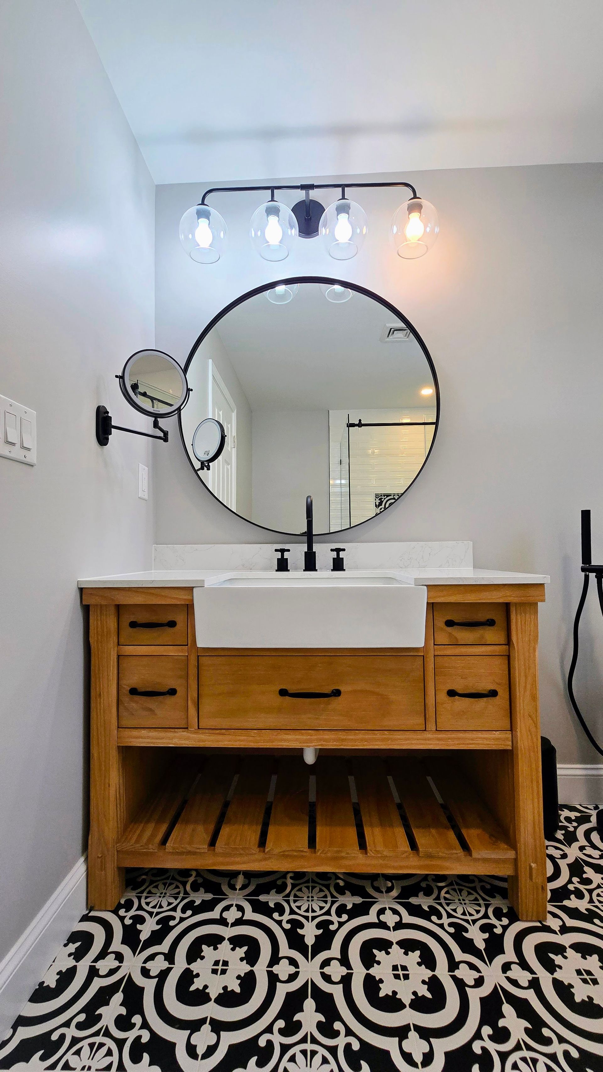A wooden bathroom vanity with a white farmhouse sink, round black-framed mirror, and patterned black and white tile floor.