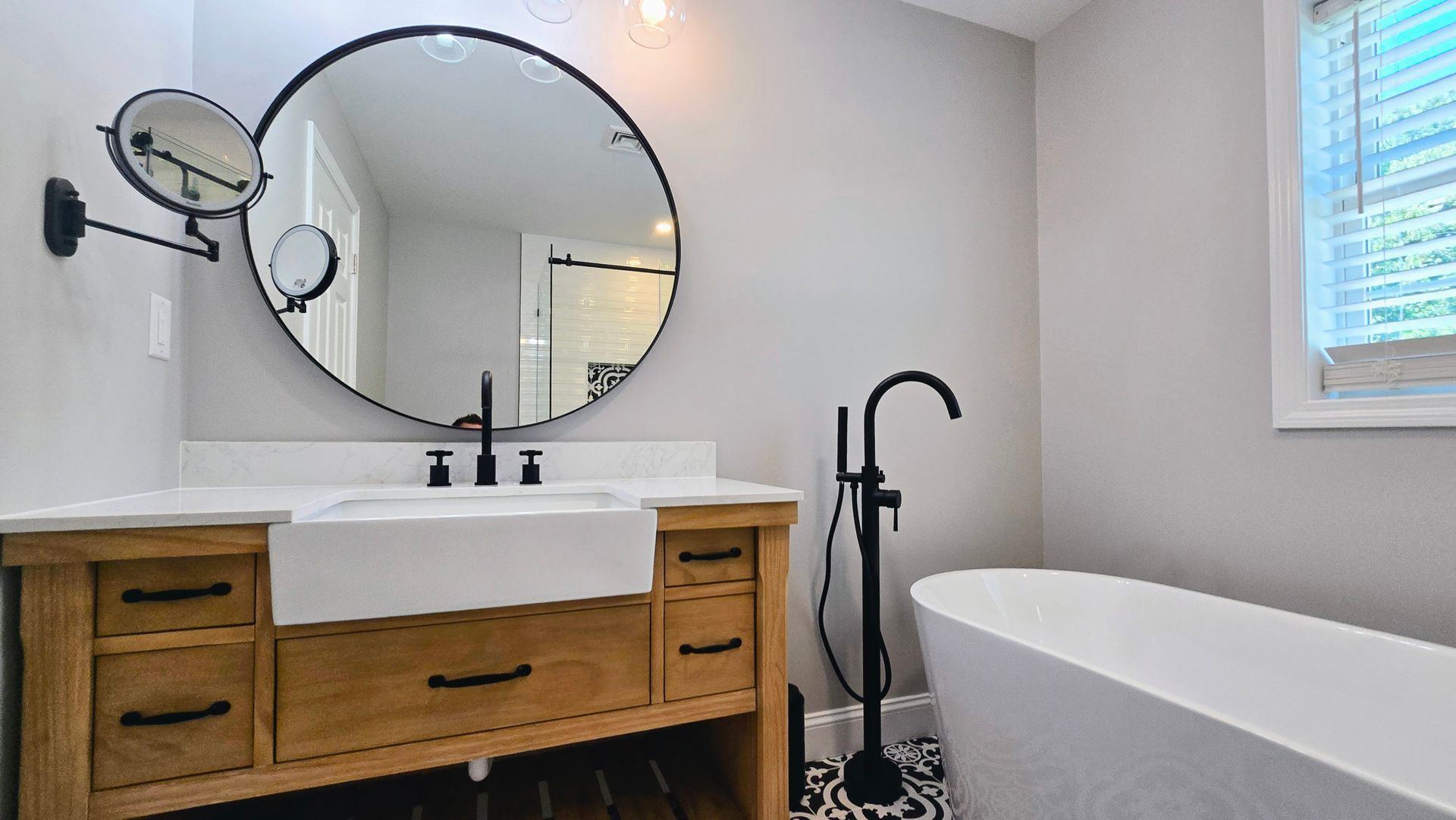 A modern bathroom renovation in Southington, CT with a wooden vanity, farmhouse sink, round mirror, and a freestanding tub against a light grey wall.