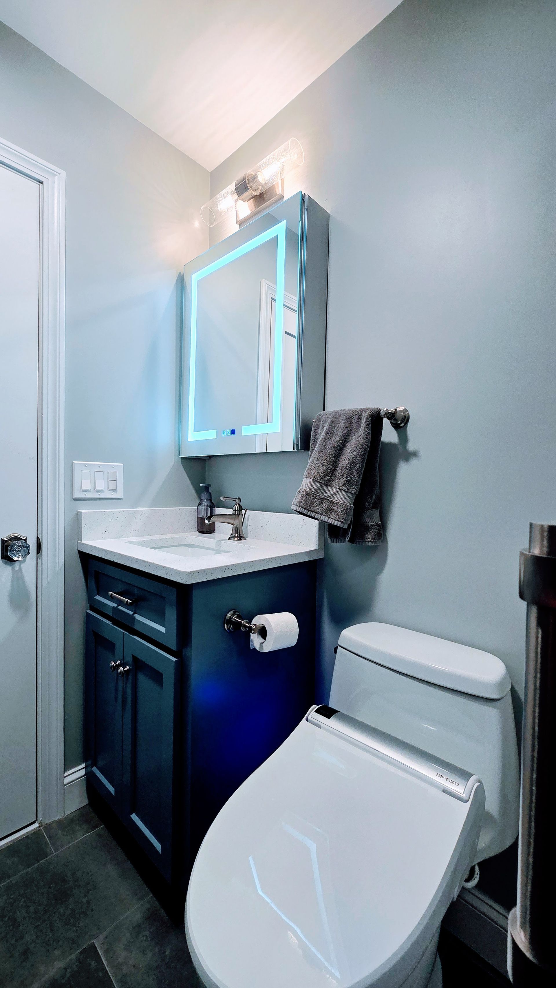Small blue West Hartford bathroom with vanity, toilet, and lit mirror. Gray walls and dark floor.