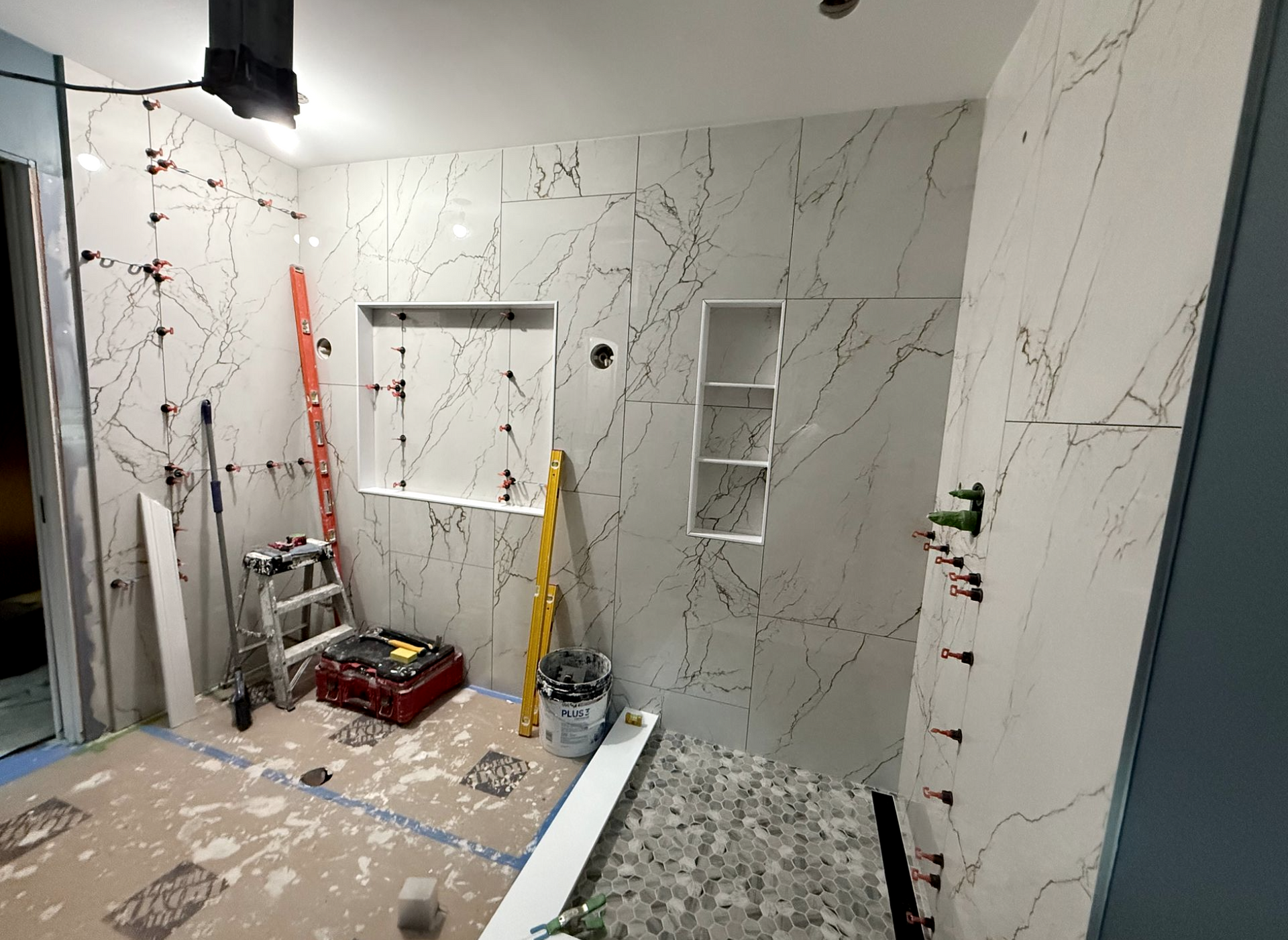 Bathroom renovation in progress; white marble-look tiles being installed, niche and shelves built in, tools and materials scattered.