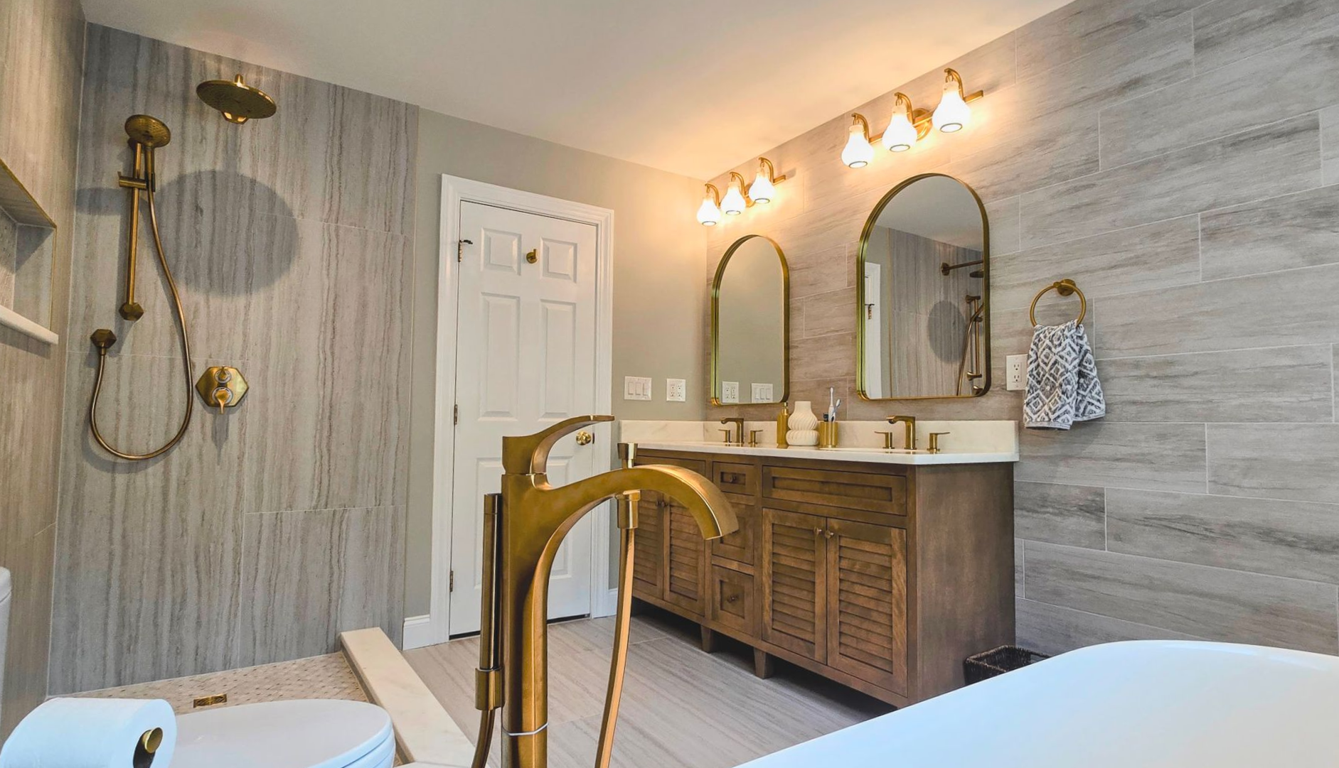 A modern bathroom in Cheshire, CT featuring a wooden vanity, gold fixtures, a rainfall shower, and light gray wood-look tiled walls.