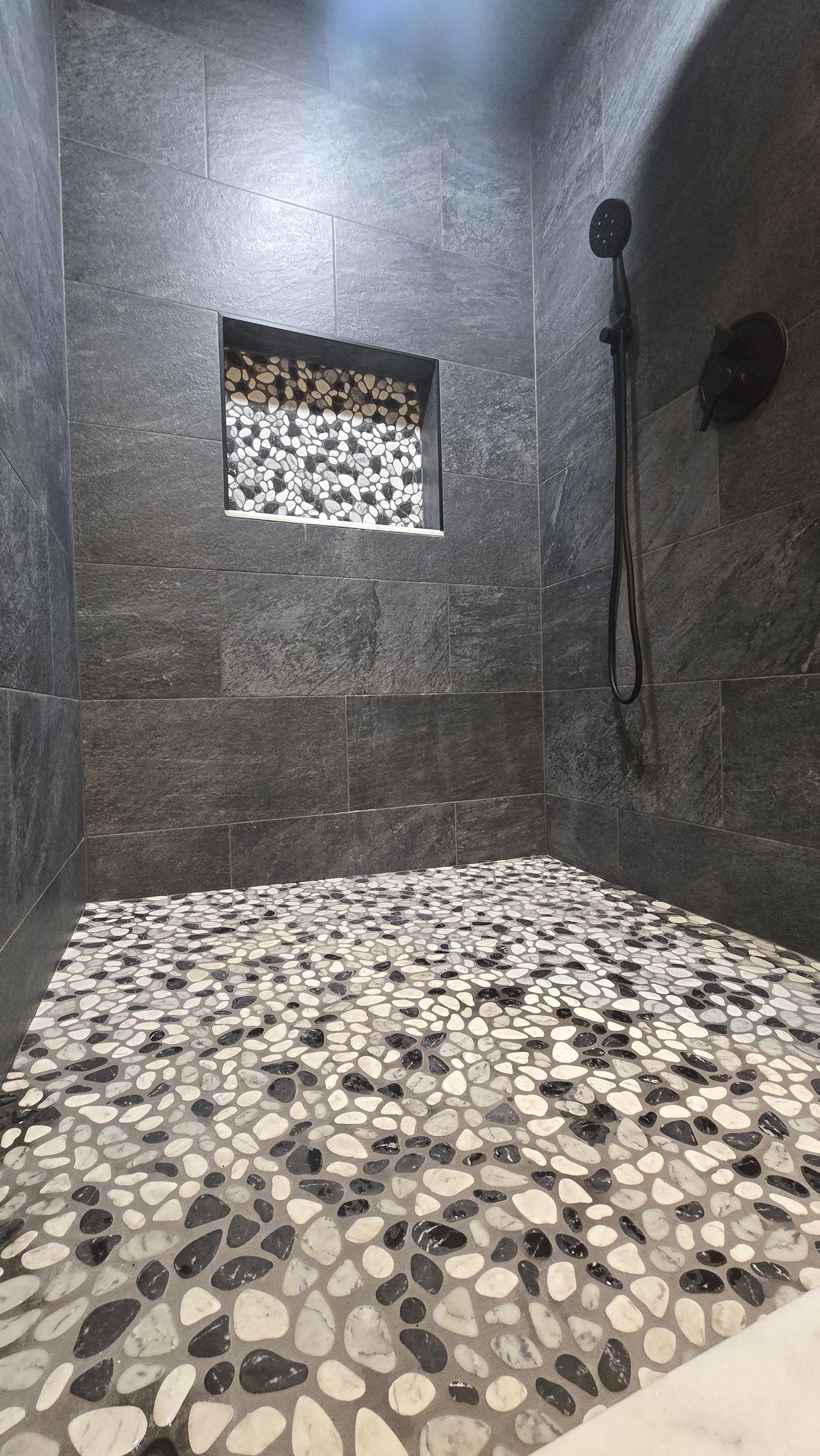 A walk-in shower with dark gray stone walls, a pebble-tiled floor, and a matching stone-tiled recessed wall niche.