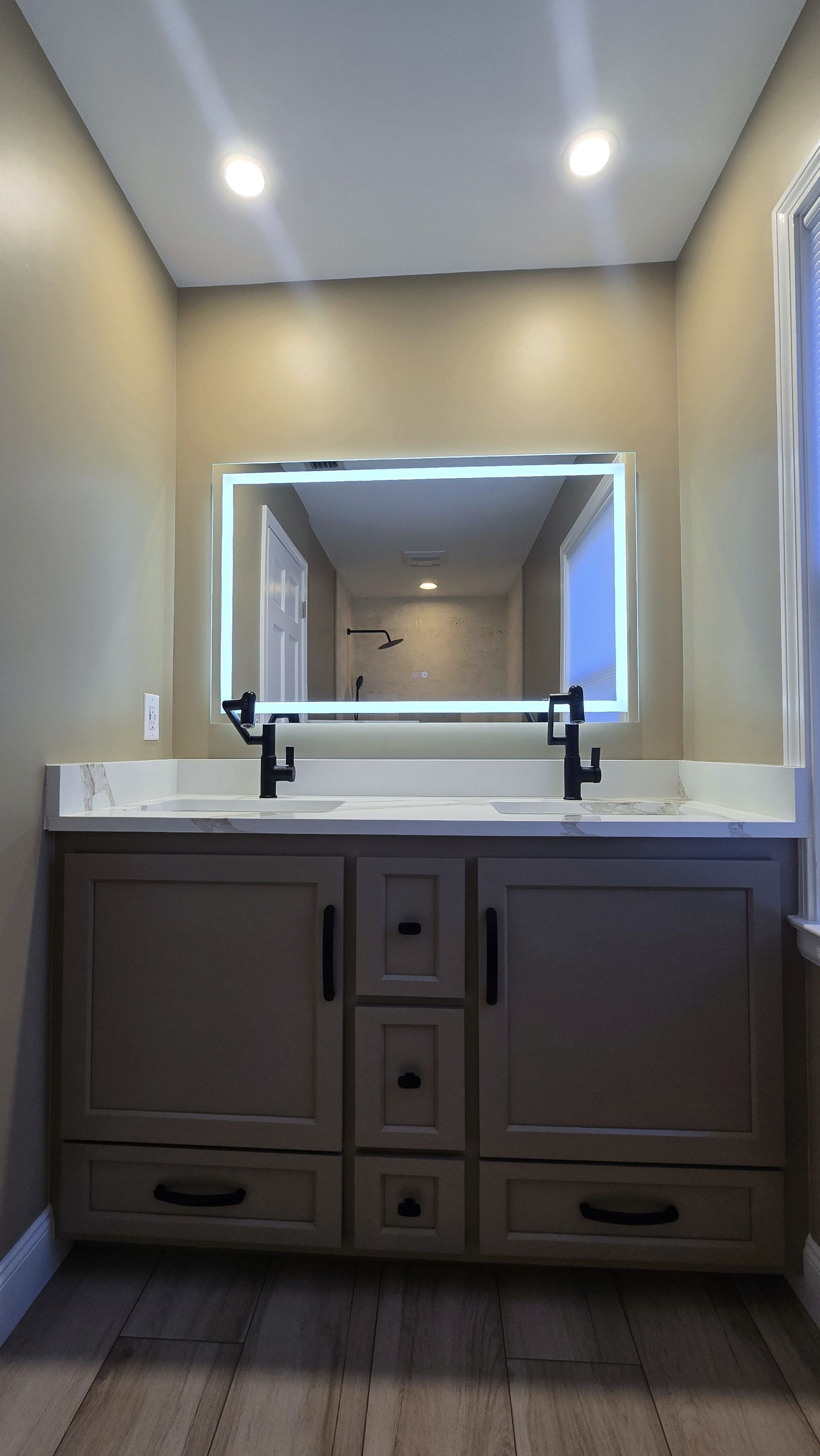 A bathroom remodel in Bristol, CT completed by First Choice Bathroom Remodeling. A vanity with backlit mirror, dark gray cabinets, white countertop, and black faucets.