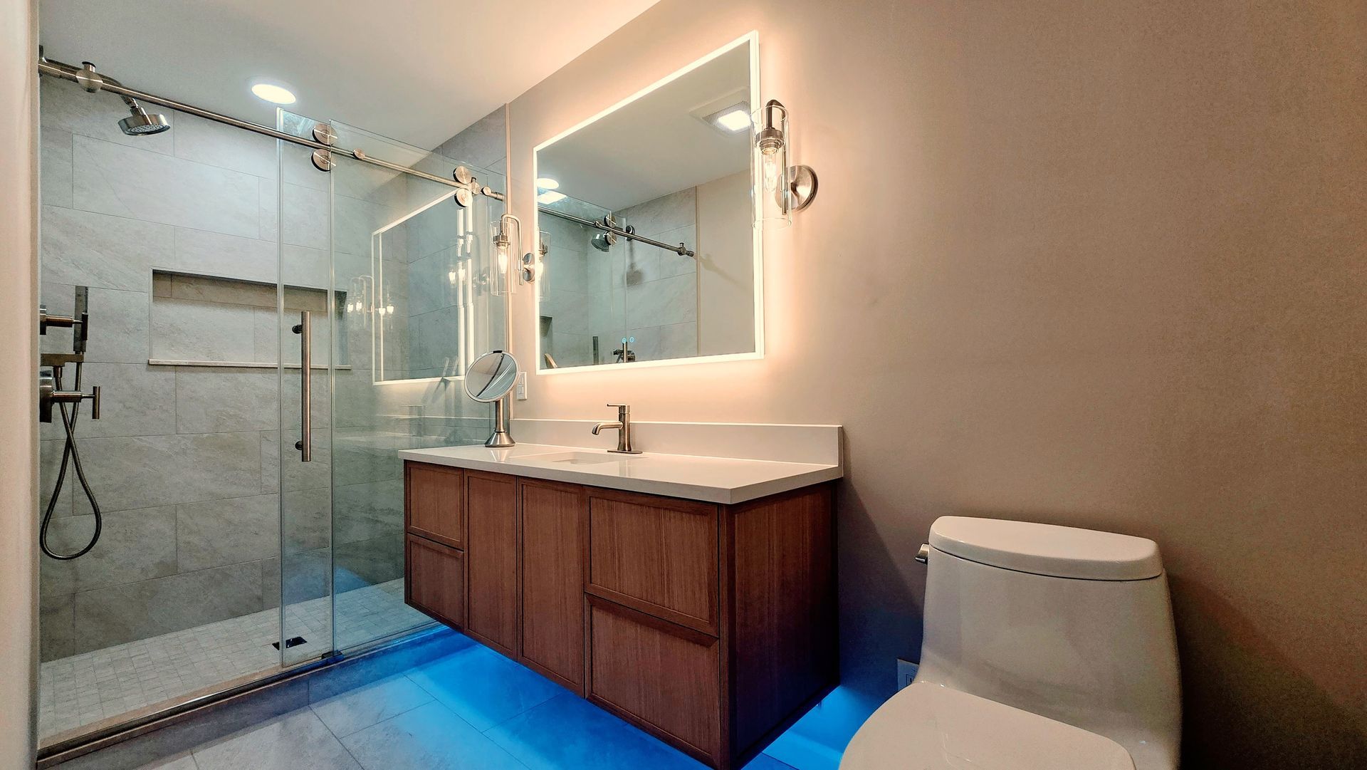 A modern bathroom in Simsbury, CT with a glass-enclosed shower, a wooden vanity with blue floor lighting, and a white toilet.