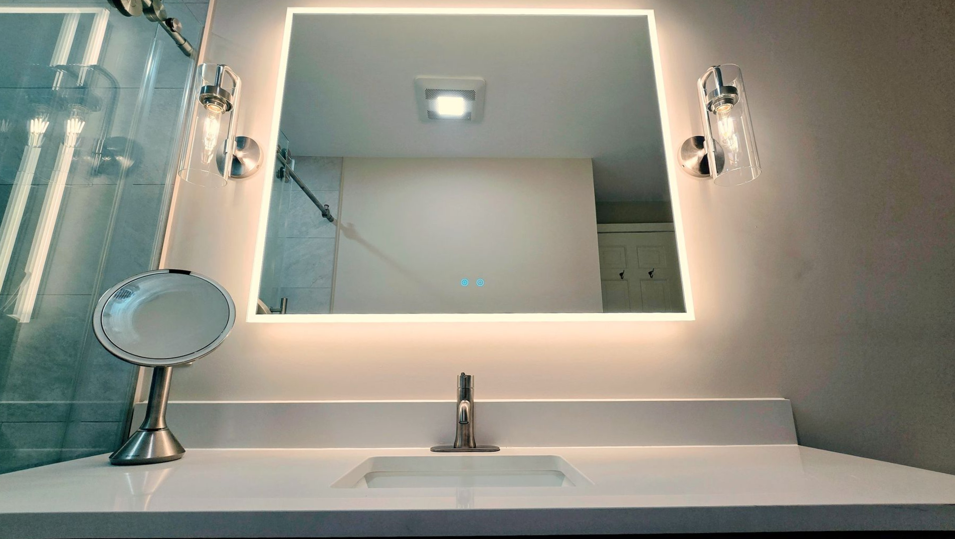 A bathroom in Simsbury, CT vanity featuring a square backlit mirror, silver wall sconces, a small round mirror, and a white countertop.