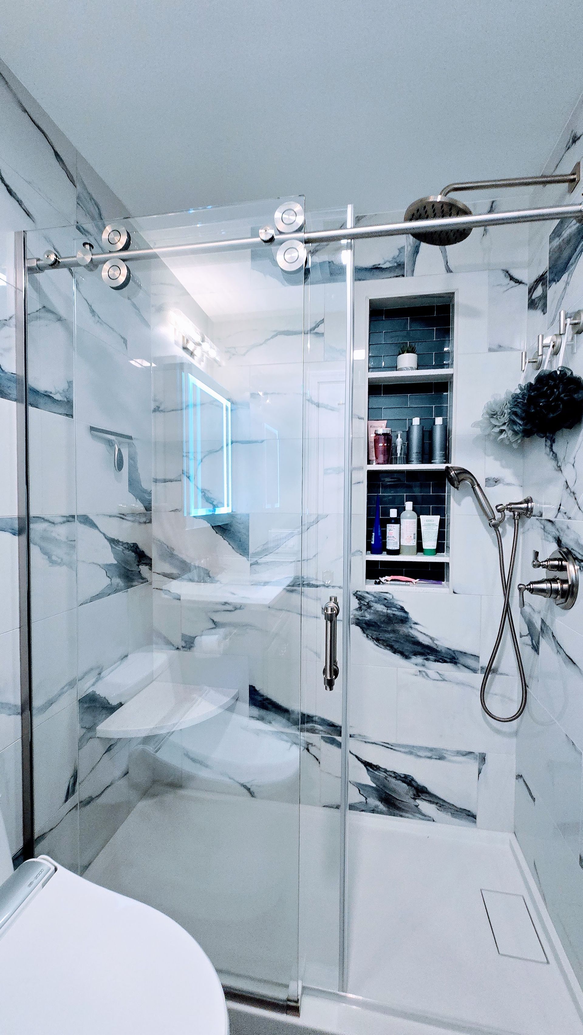 Modern bathroom with marble walls, glass shower door, and built-in shelving with toiletries.