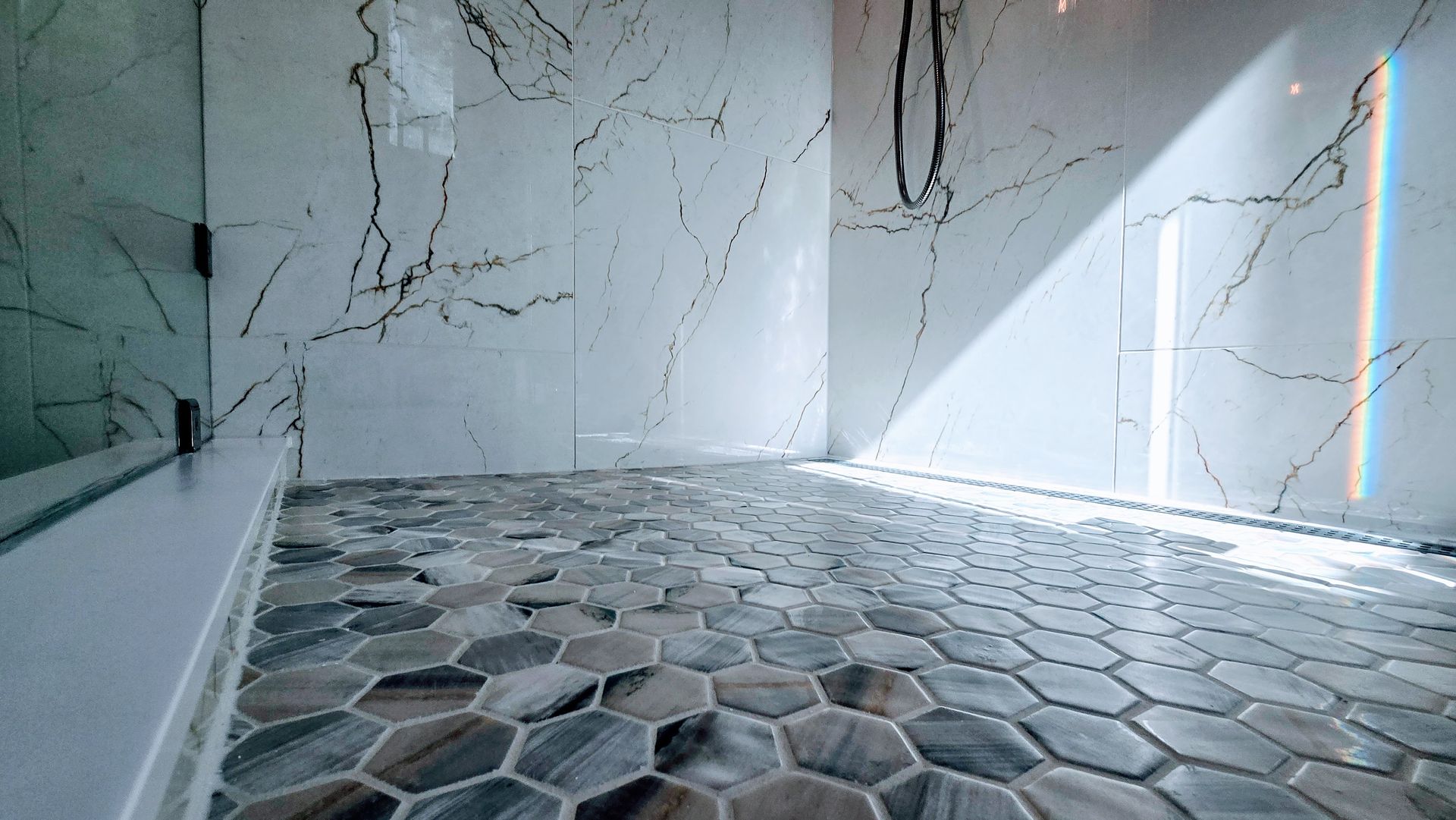 Shower with marble walls, hexagon tile floor, and glass door. Sunlight shines through.