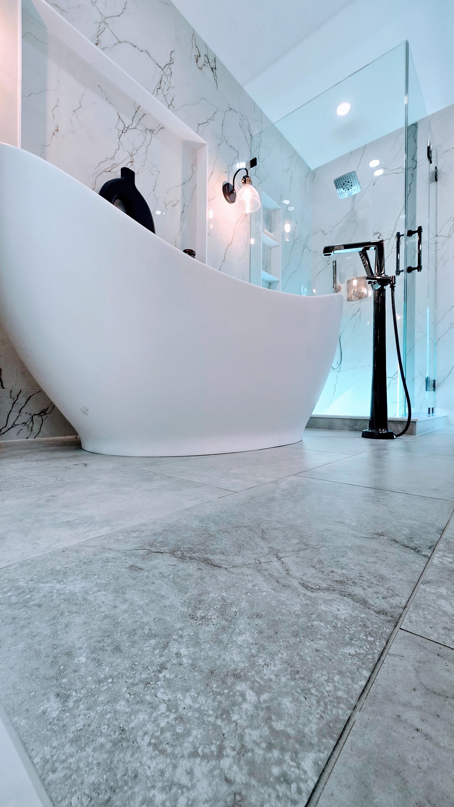 White oval bathtub in a modern bathroom with grey tiled floor and marble walls.