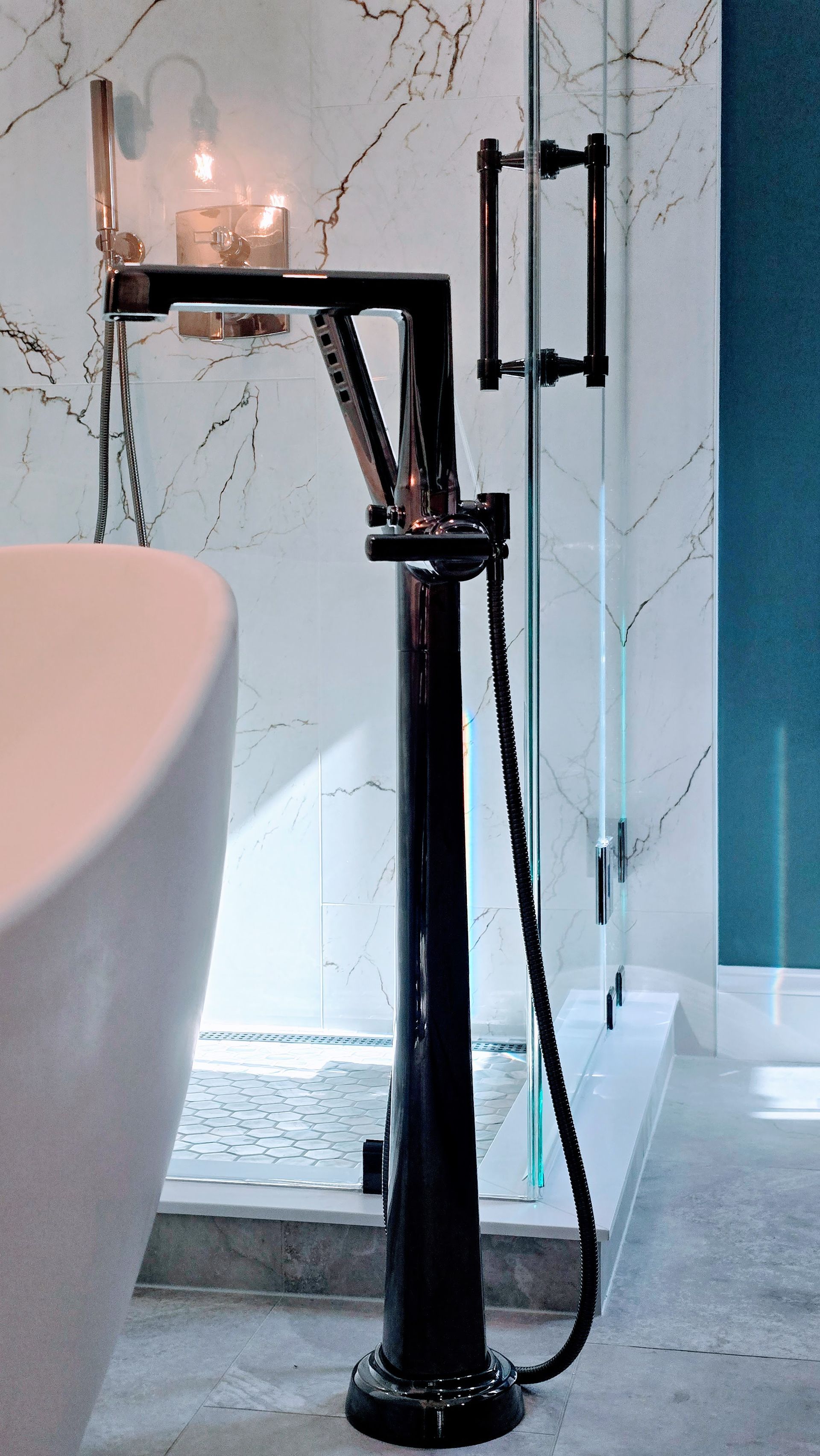 Black floor-mounted bathtub faucet with hand shower near a white tub and shower door.