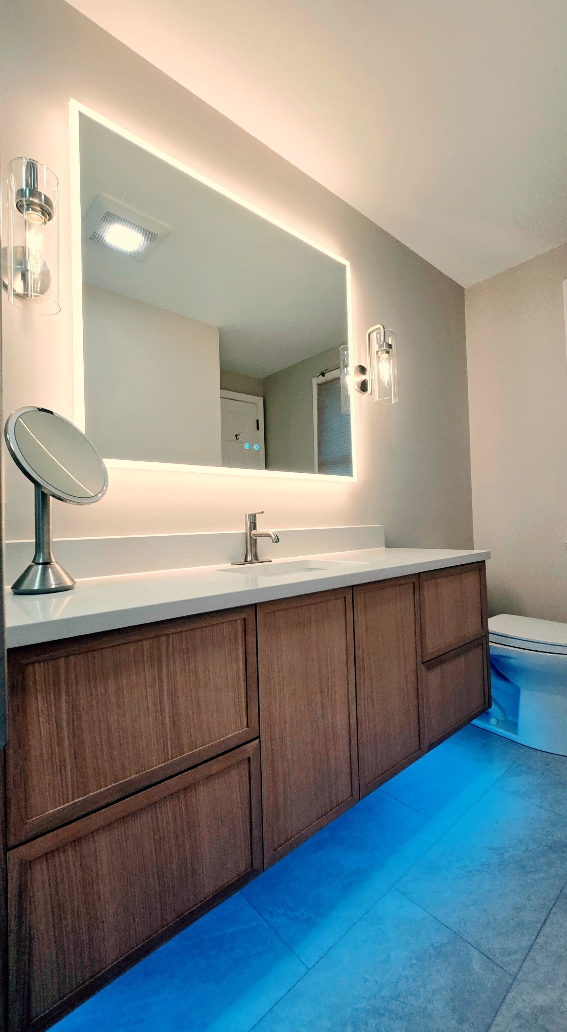 A wooden vanity with a backlit mirror, a standing mirror, and blue floor lighting in a modern bathroom.