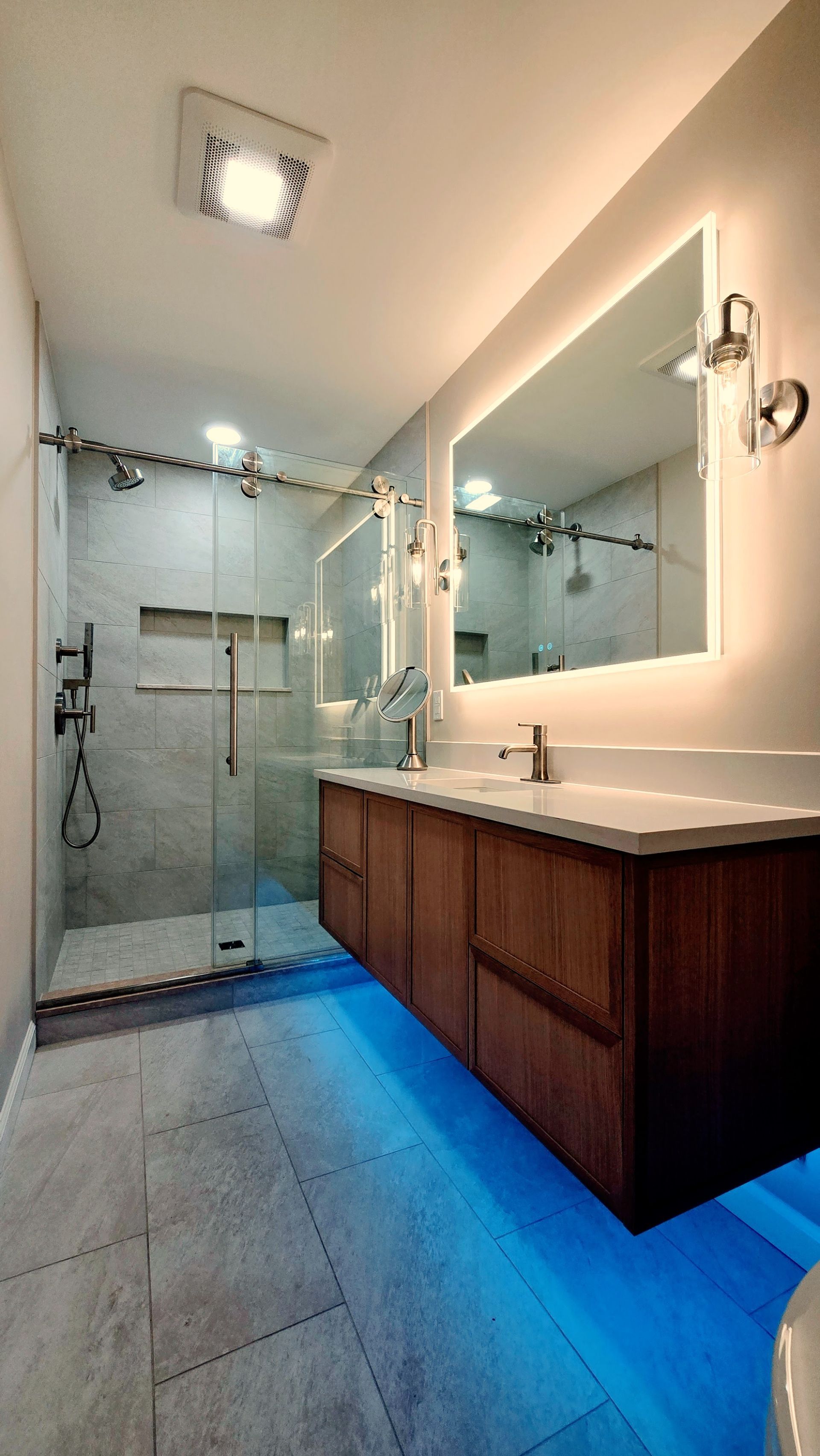 A modern bathroom in Simsbury, CT with a glass-enclosed shower, a dark wood vanity with a backlit mirror, and blue floor accent lighting.
